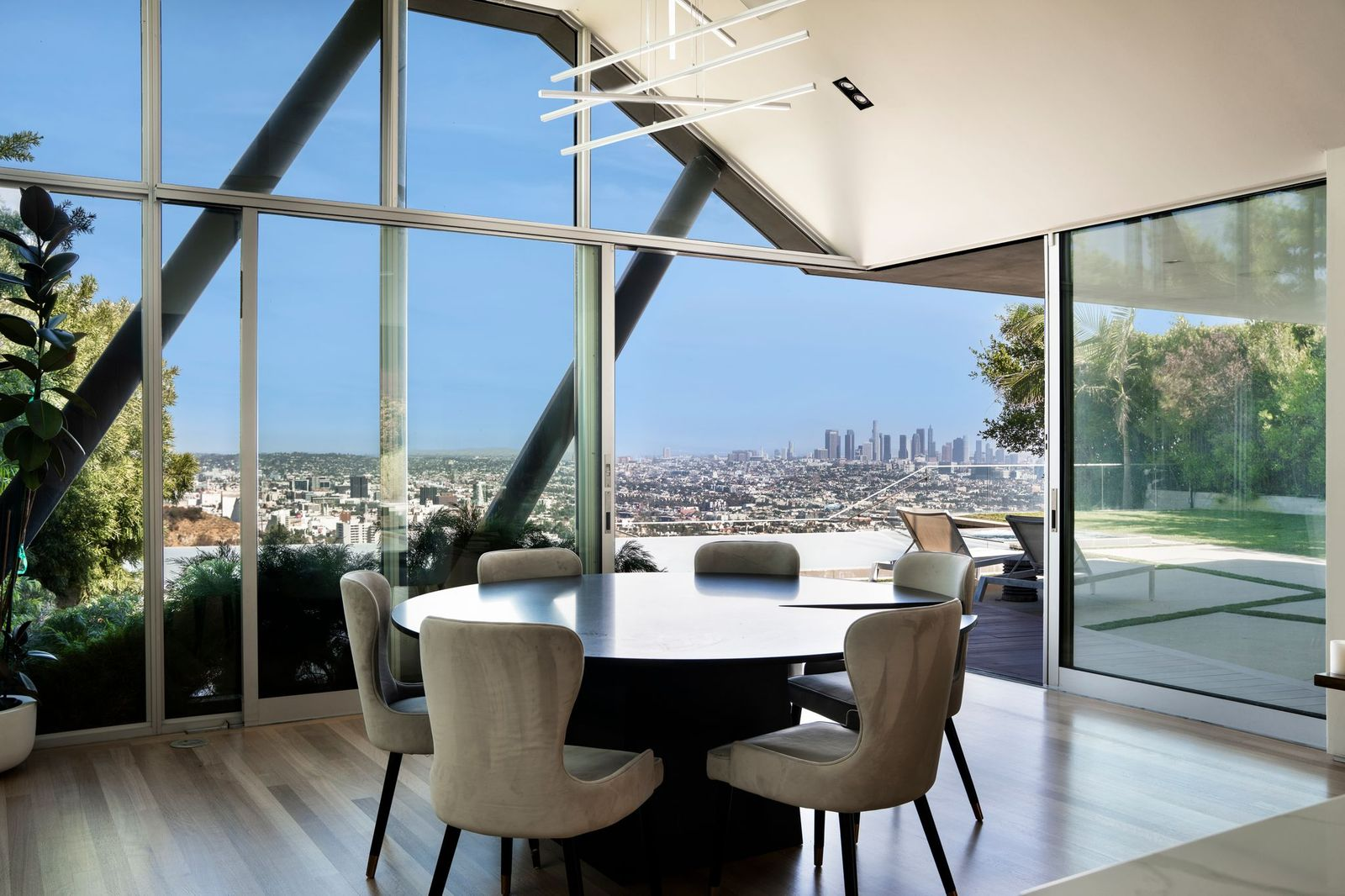 A round dining table sits beneath a contemporary linear chandelier, surrounded by upholstered chairs and framed on two sides by floor-to-ceiling glass walls that overlook the backyard and the Los Angeles skyline.