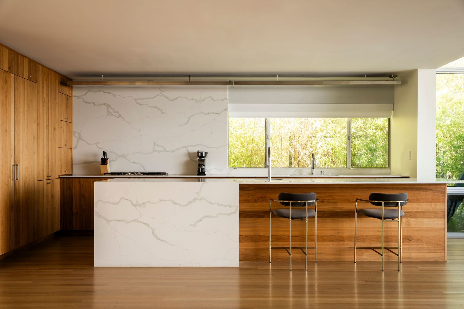 A waterfall-edge island at the front of the space combines white stone surfaces with warm wood paneling and bar seating positioned along one side for casual dining.