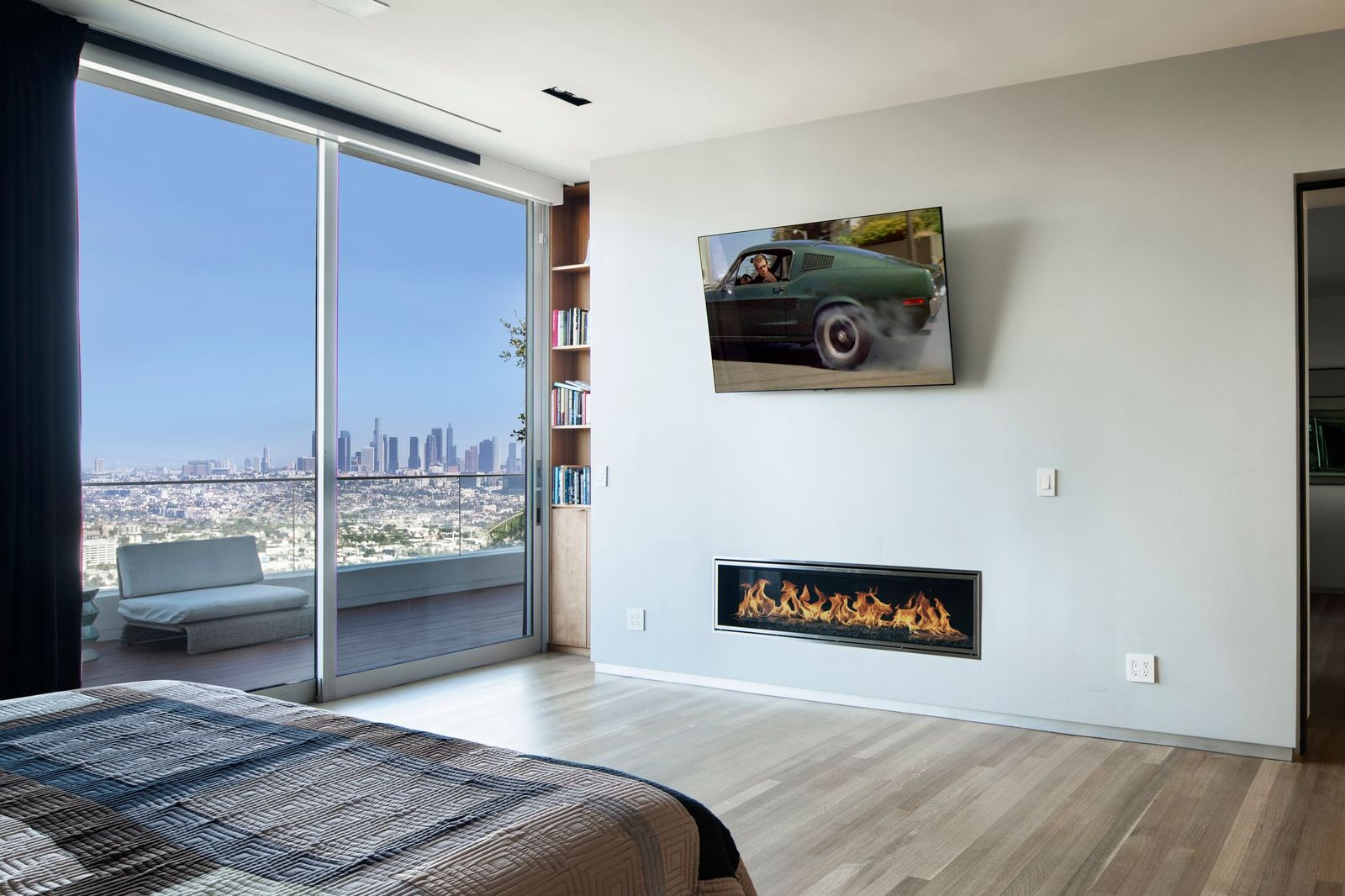 The bedroom is intentionally minimalist in its design, with wide-plank wood floors running underfoot, while a sleek linear fireplace is integrated into one wall beneath a mounted television.