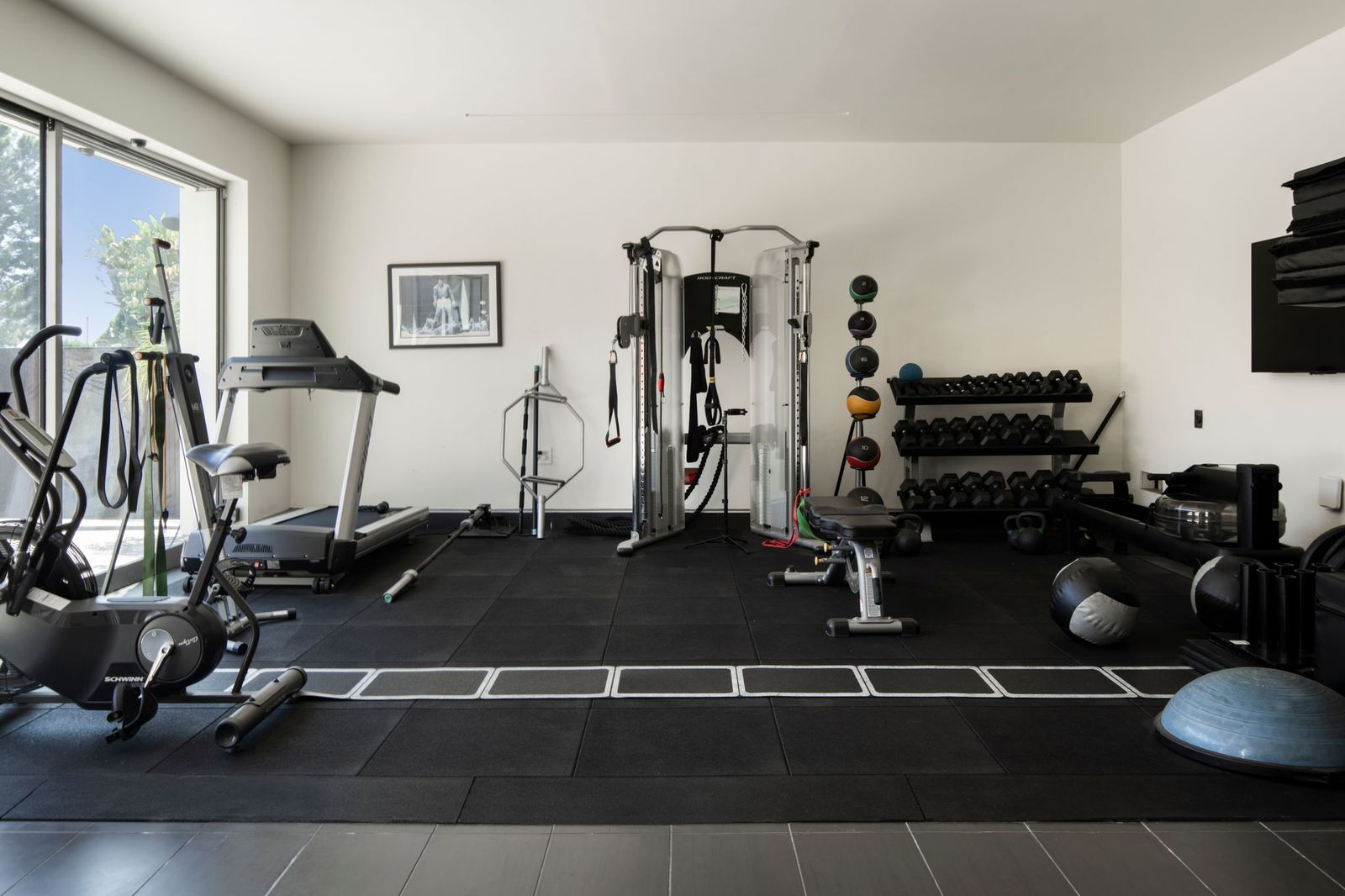 Along one side are cardio machines, including a treadmill and a stationary bike, while the center of the room features a cable machine and an adjustable weight bench.