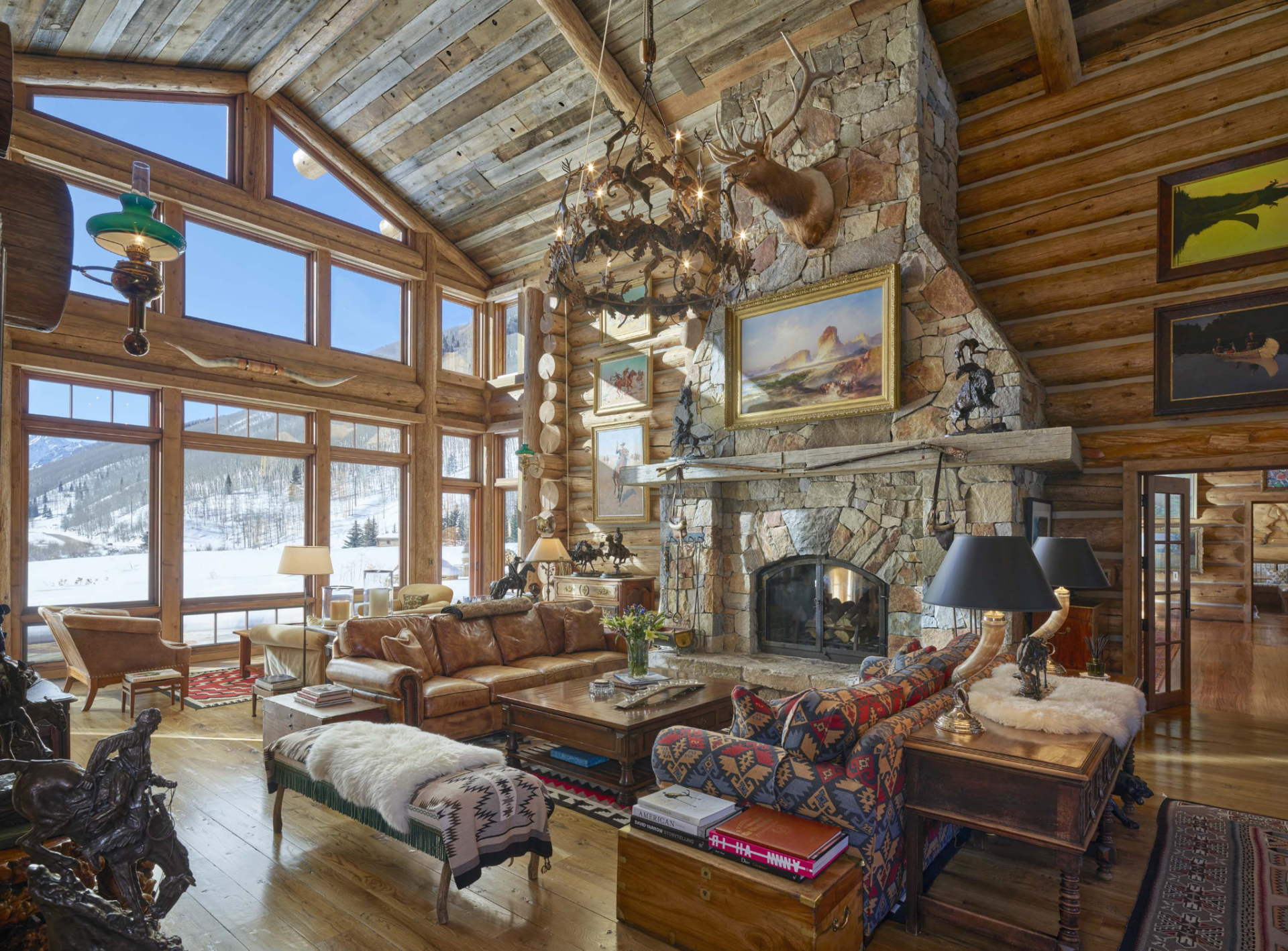 At the center of the room, a monumental stone fireplace rises to the ceiling, anchored by a timber mantel and surrounded by Western artwork, sculptures, and collected memorabilia. 