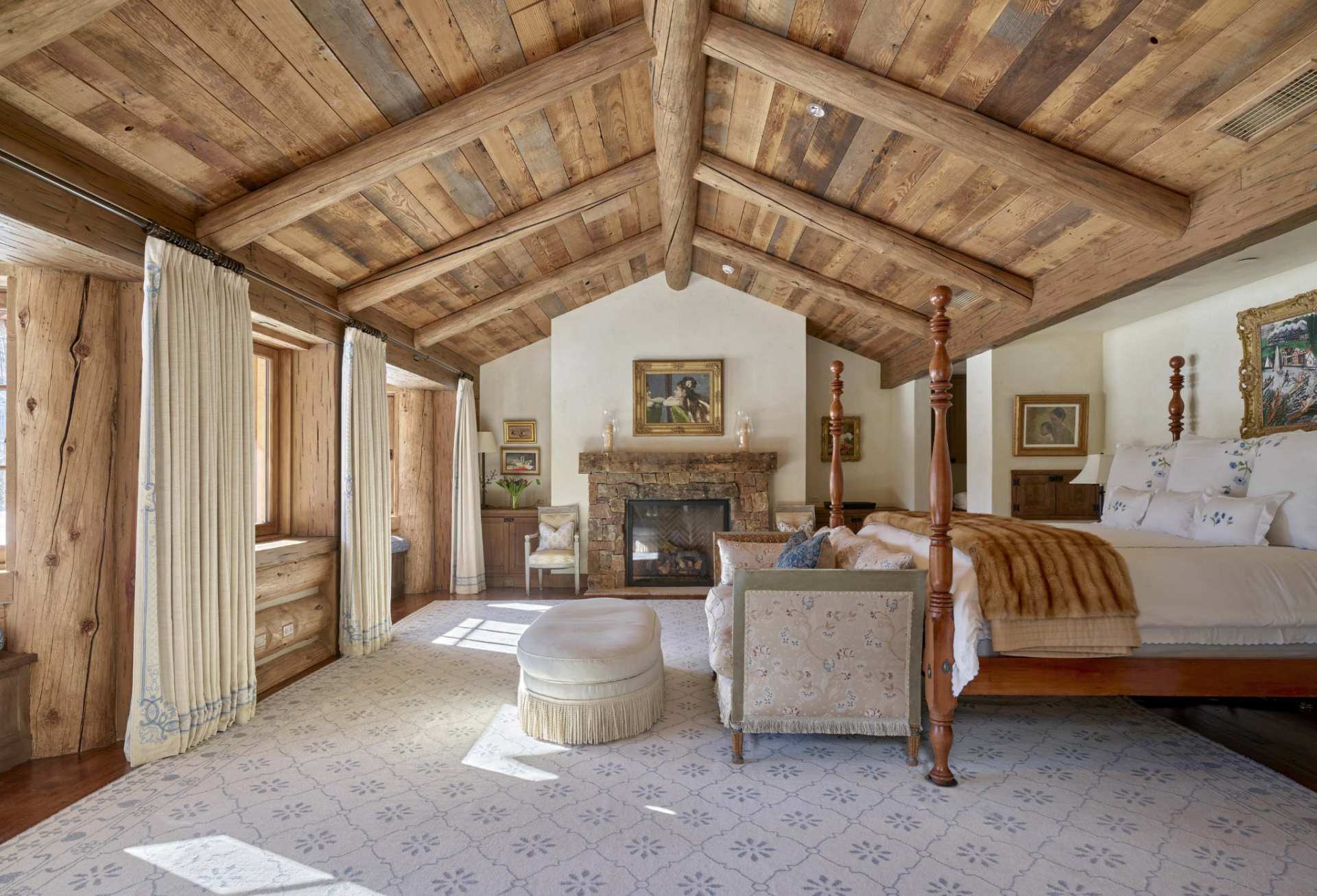 A four-poster bed sits in the center of the room, positioned opposite a stone fireplace and a wall of windows.