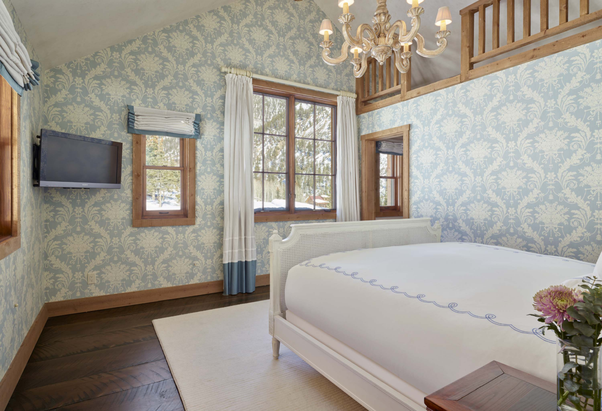 Another guest bedroom introduces a lighter, more feminine aesthetic through pale blue patterned wallpaper, white furnishings, and a more traditional cottage-inspired design. 