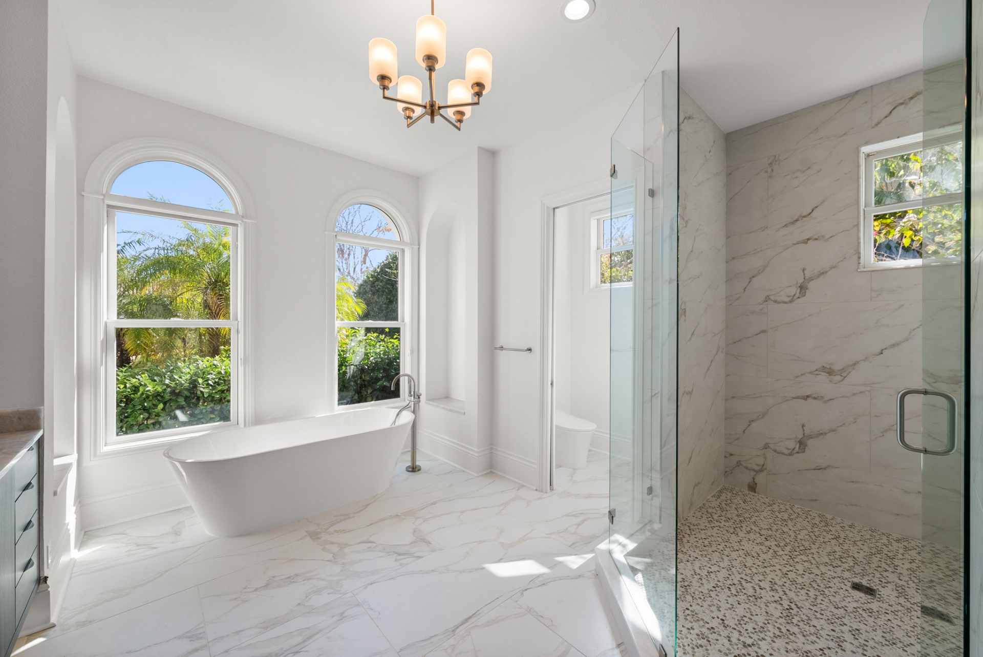 At the opposite end of the room, a freestanding soaking tub is positioned beneath arched windows, while a large glass-enclosed shower finished in marble tile completes the neutral space.