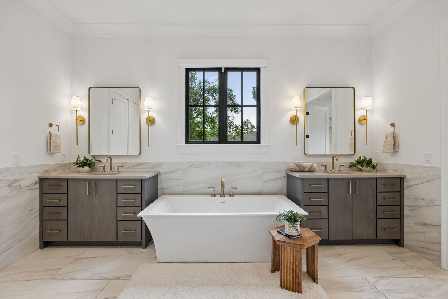 Centered beneath a large window, a freestanding soaking tub is framed by matching dual vanities finished with gray cabinetry and brass hardware.
