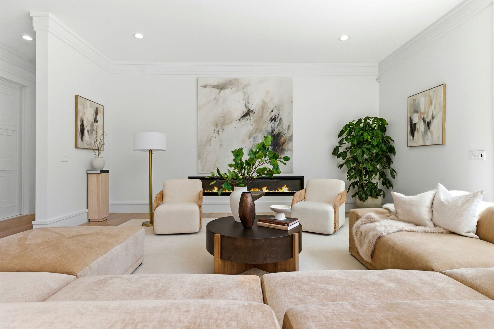 Off to the left, the main living room features a long linear fireplace set beneath oversized abstract artwork, with a collection of neutral-toned seating arranged around a circular wood coffee table.