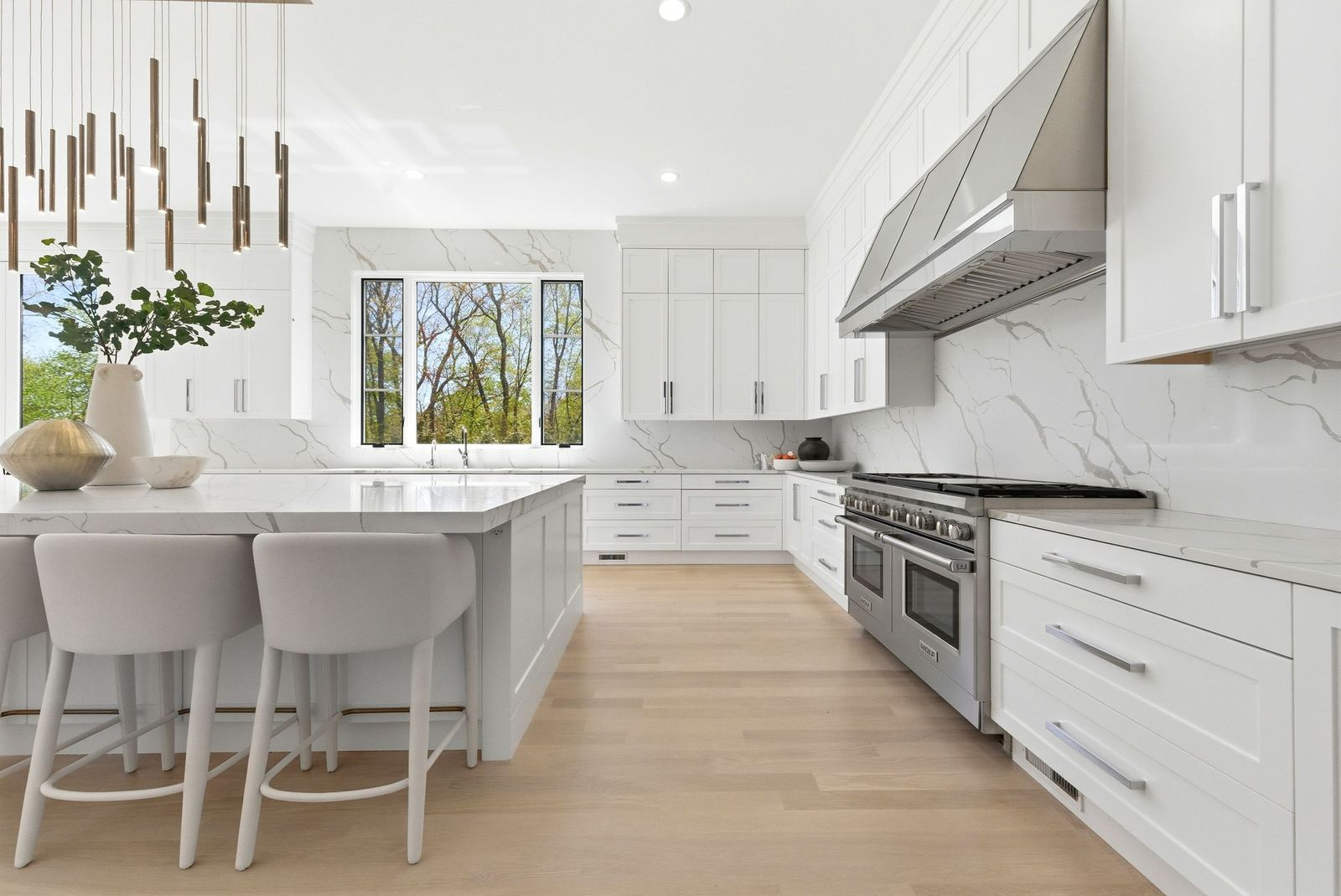 A large center island provides seating for several guests beneath a modern pendant fixture, while professional-grade appliances, including a range and oversized vent hood, are integrated into the perimeter cabinetry.