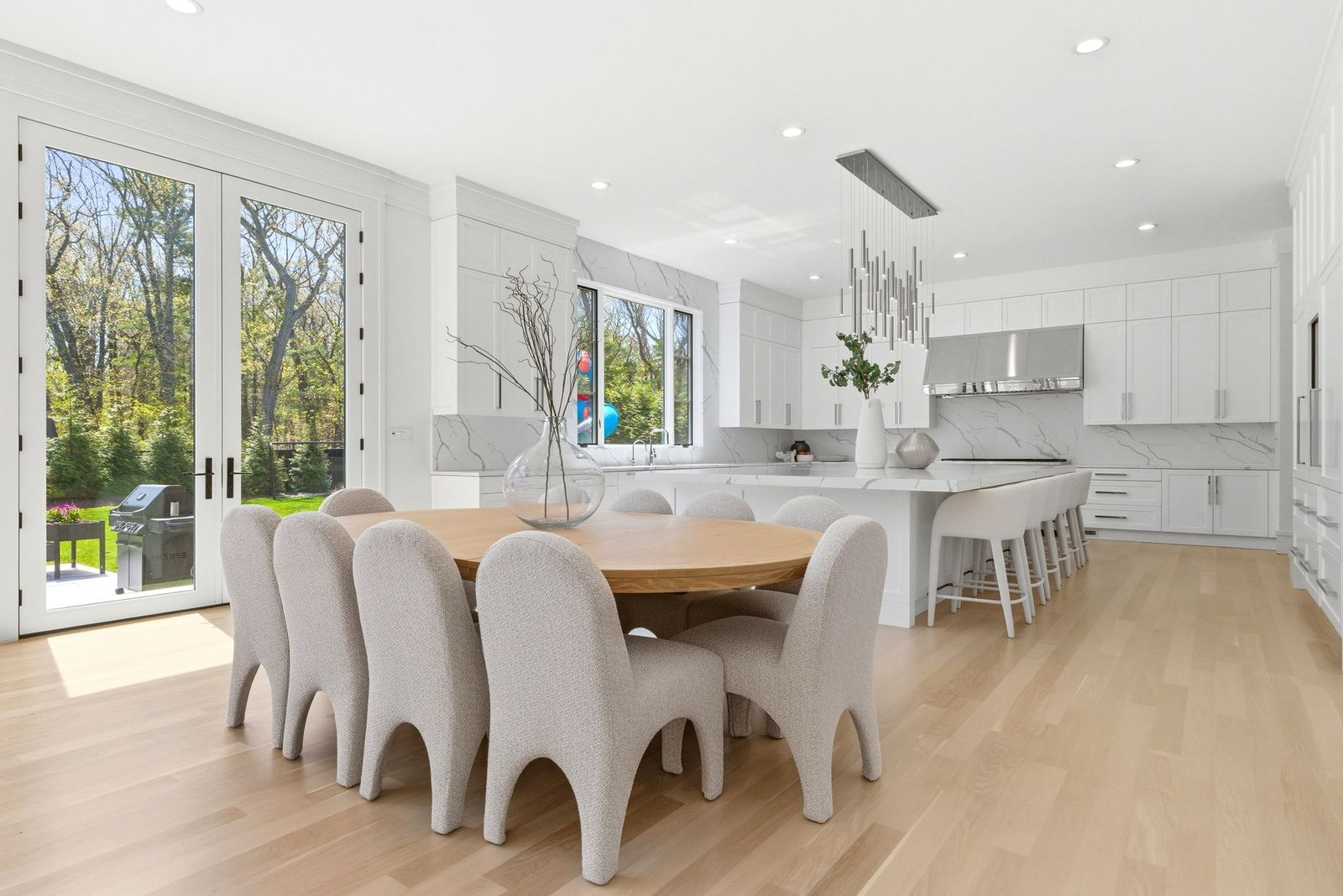 A round wood table surrounded by sculptural upholstered chairs sits beside French doors leading to the backyard, creating an informal space for everyday meals.