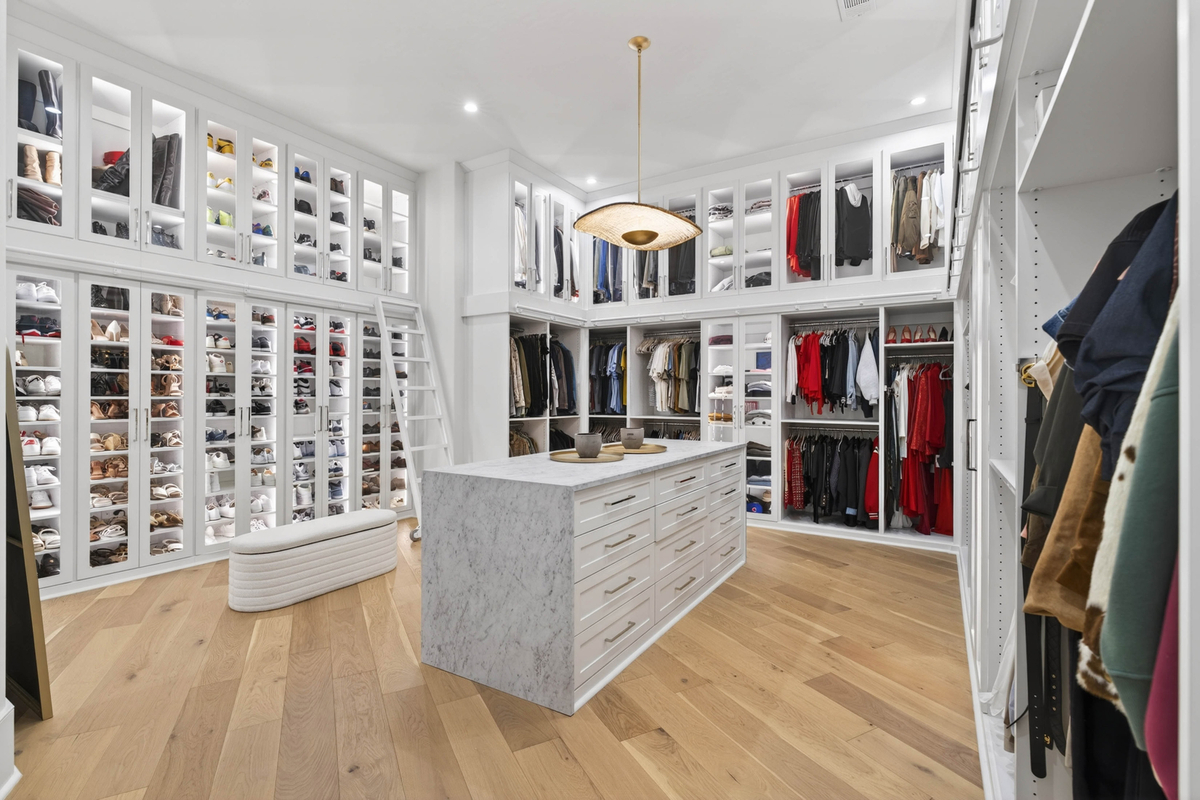 Connected to the suite is an enormous dressing room with glass-front display cases showcasing shoes, handbags, and accessories alongside hanging storage and shelving.