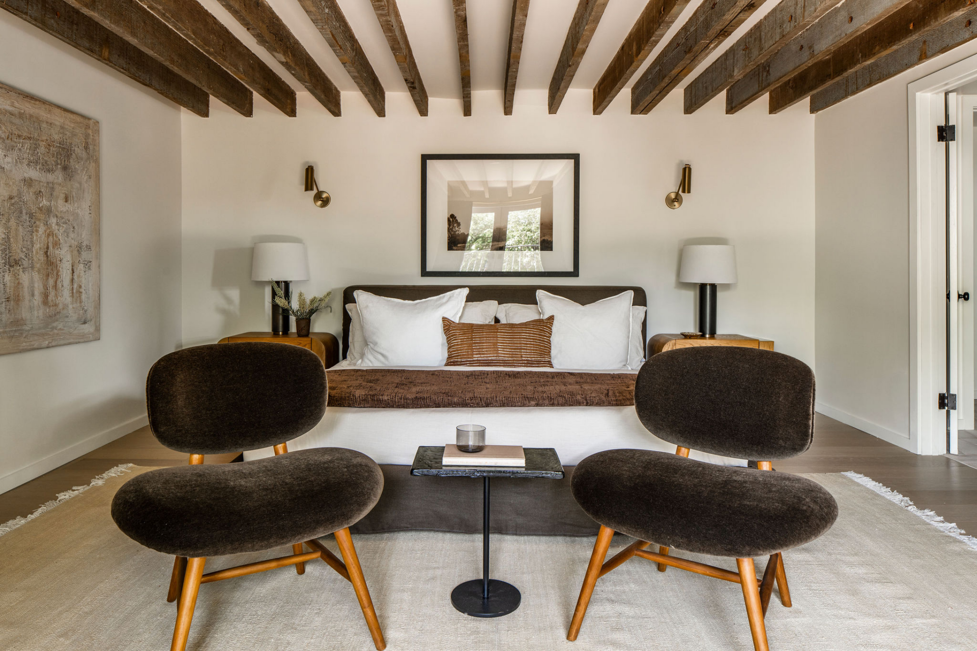 Inside the bedroom, a pair of sculptural lounge chairs create a dedicated sitting area at the foot of the plush bed, while soft textures and natural materials keep the space simple and inviting. 