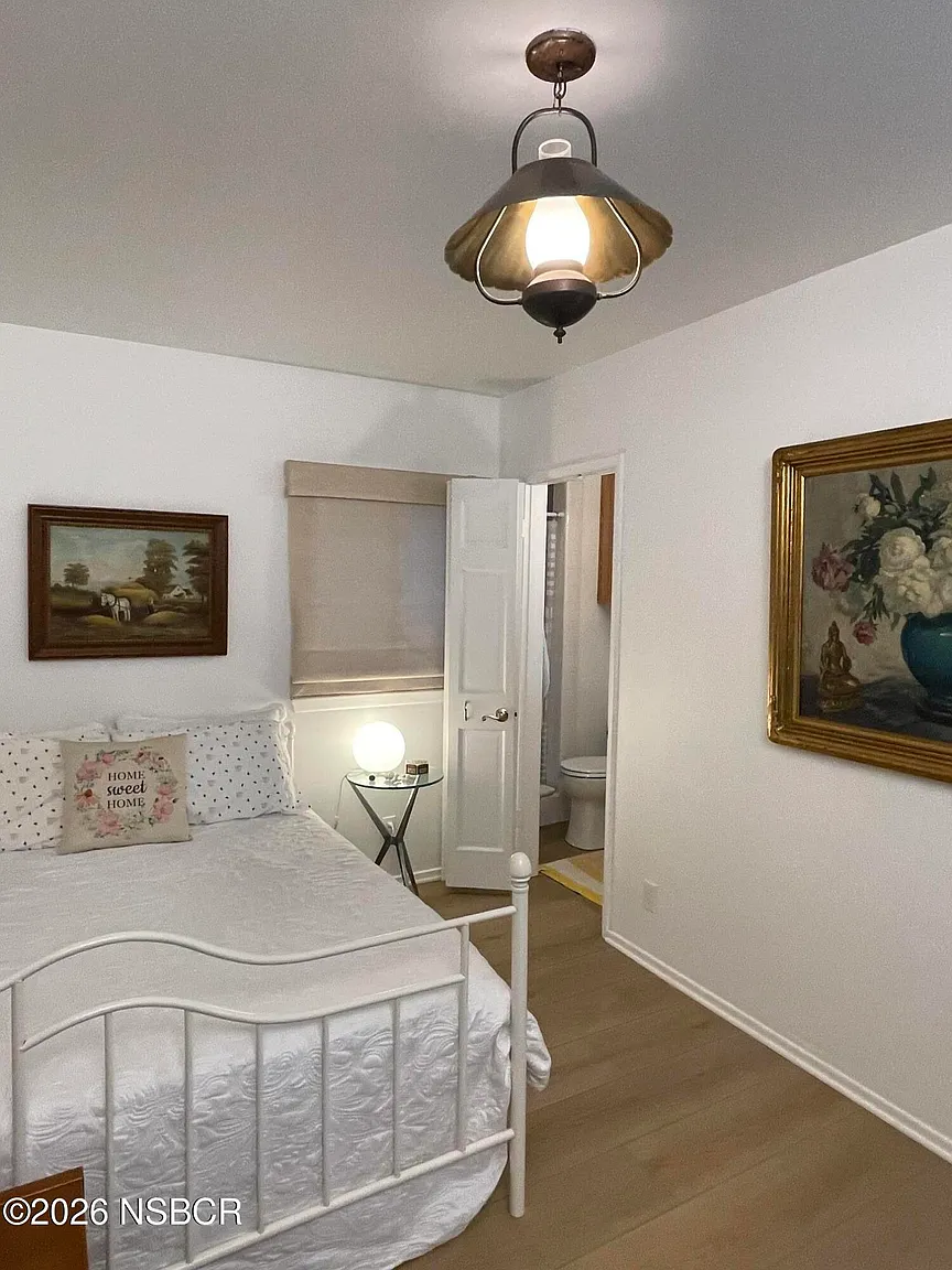 The first bedroom has a cottage-like feel, centered around a white metal bed frame dressed in crisp white bedding, with a connected en-suite bathroom.