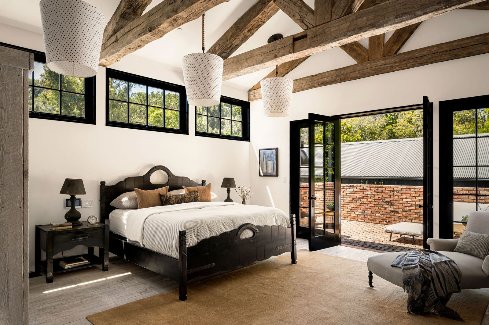 The bedroom centers around a dark wood bed positioned beneath vaulted ceilings crossed by massive reclaimed beams.