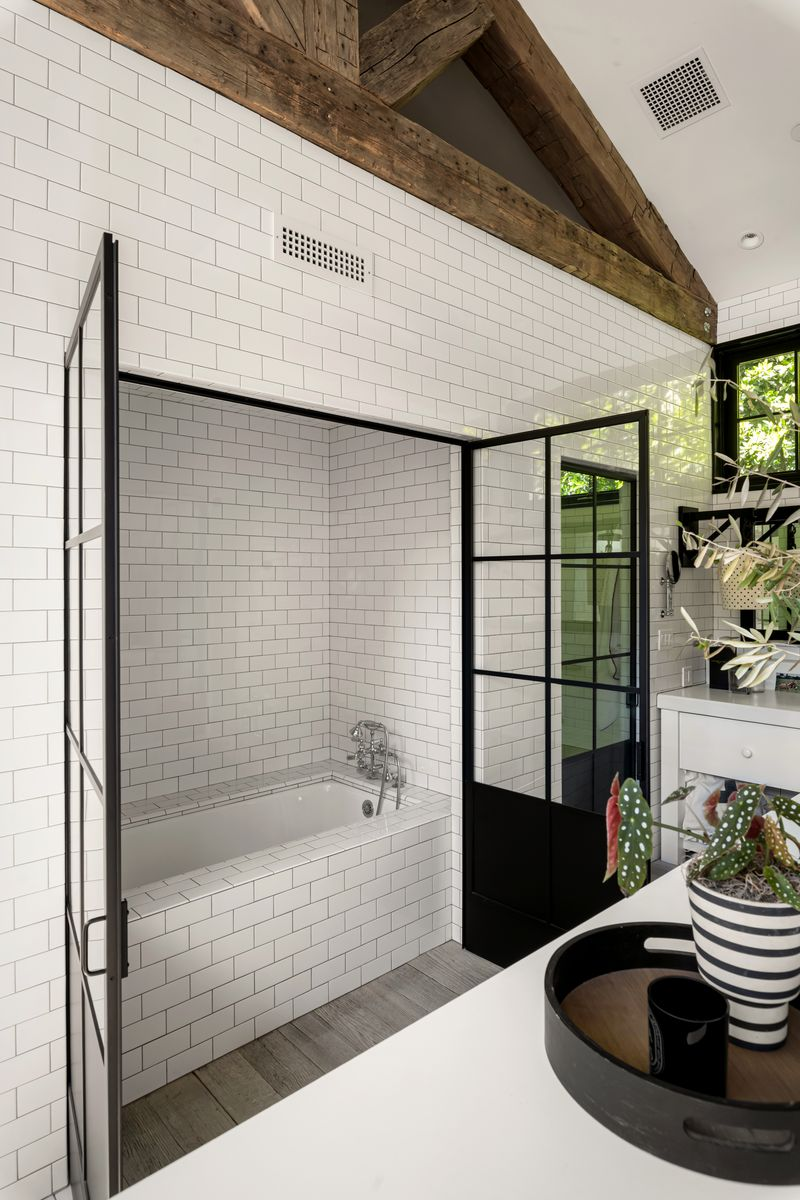 The entire room is surrounded by walls of white subway tile, contrasted by black steel-framed doors that open up to a soaking tub and walk-in shower.