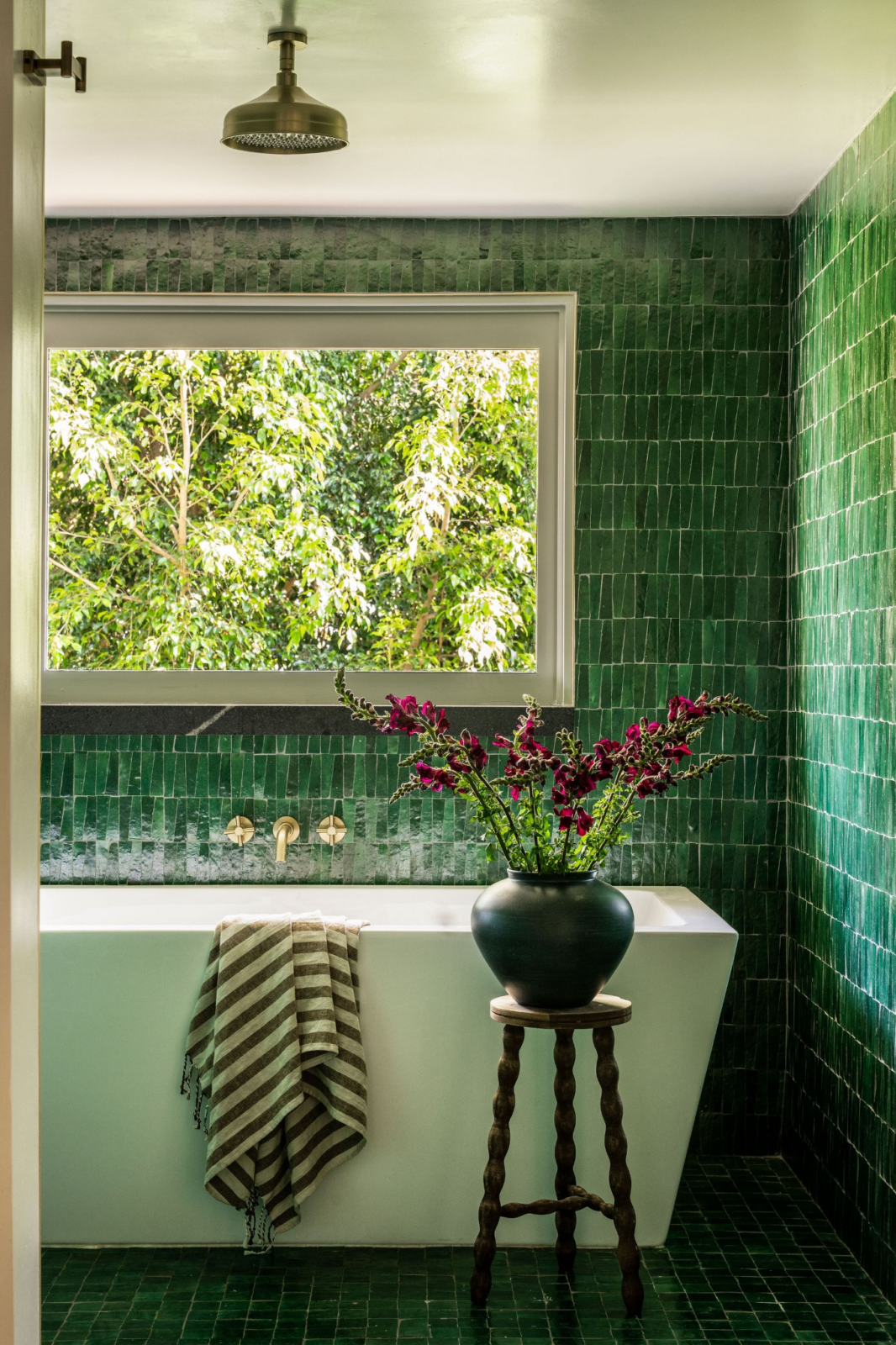 In the bathing area, deep green tile wraps the walls and floors around a freestanding soaking tub positioned beneath a large picture window.
