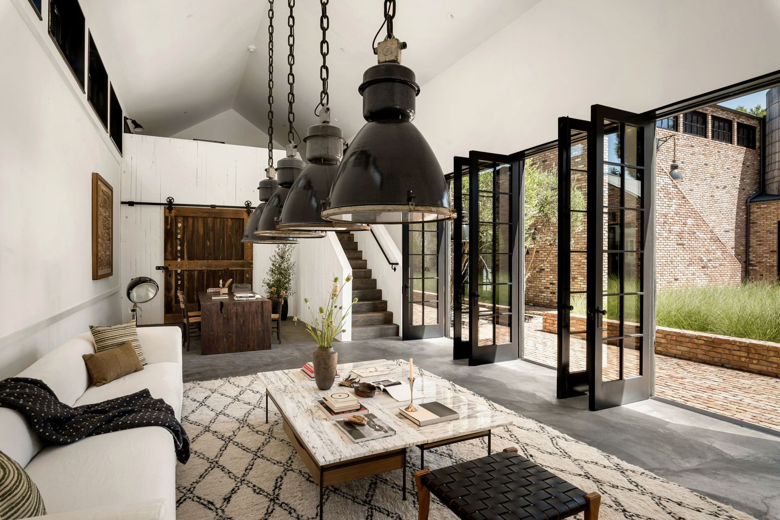 The main level features soaring ceilings, oversized industrial pendant lights, polished concrete floors, and a wall of steel-framed doors that open directly to a brick courtyard.