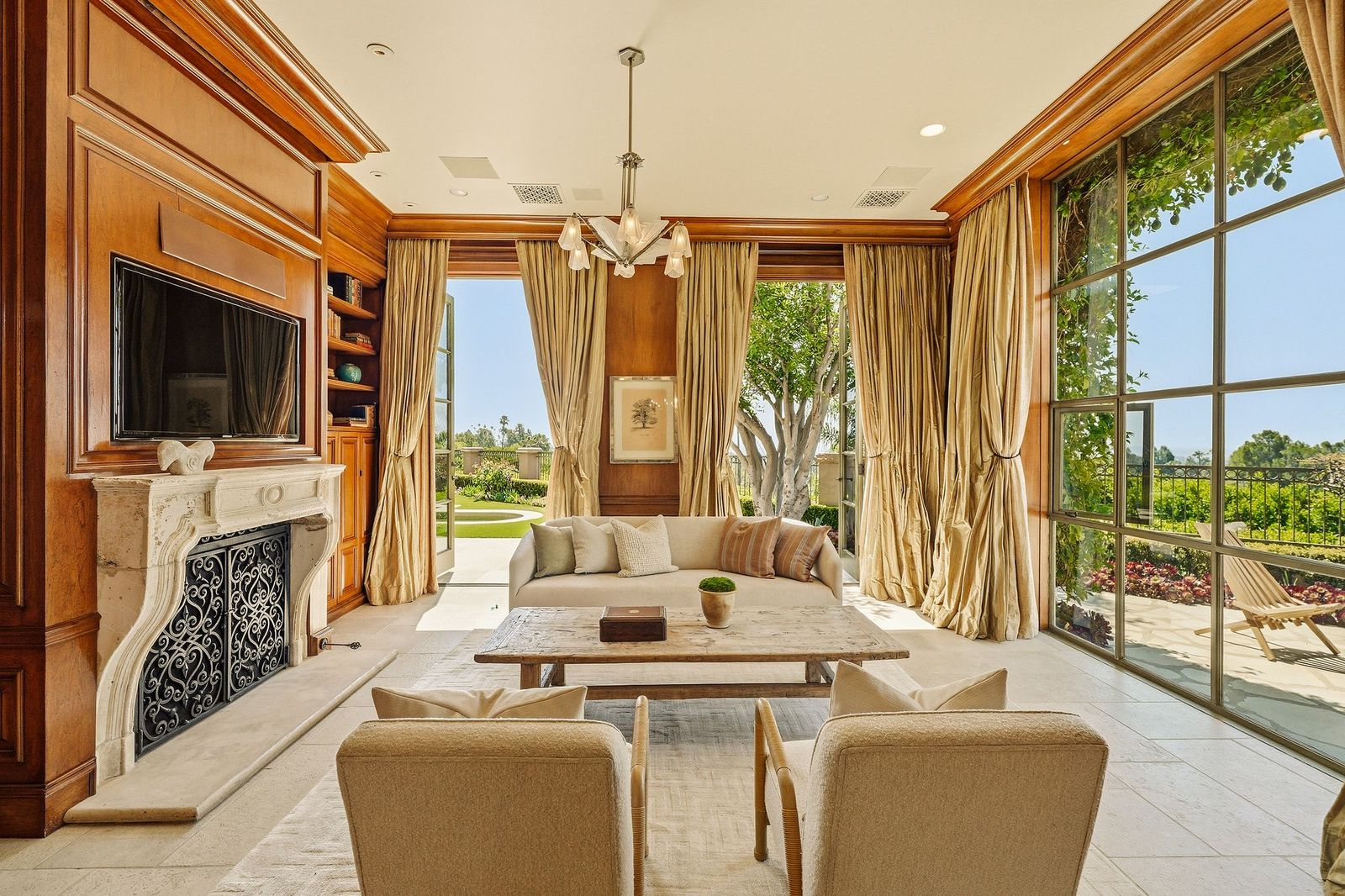 Tall steel-framed windows and French doors wrap two sides of the room and draw in natural light, while a wall-mounted television is set above the fireplace.