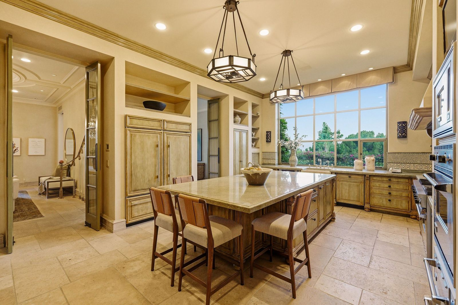 The chef's kitchen features custom distressed cabinetry and expansive stone countertops arranged around a large center island with bartop seating. 