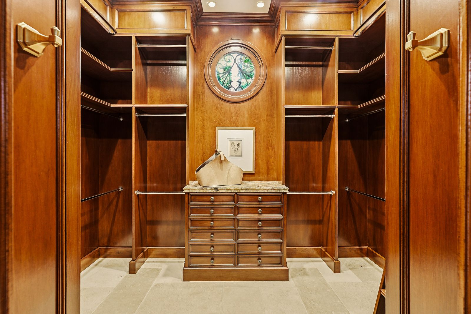 Walnut cabinetry with hanging space, shelving, and banks of drawers arranged around a central storage island beneath a round leaded-glass window. 