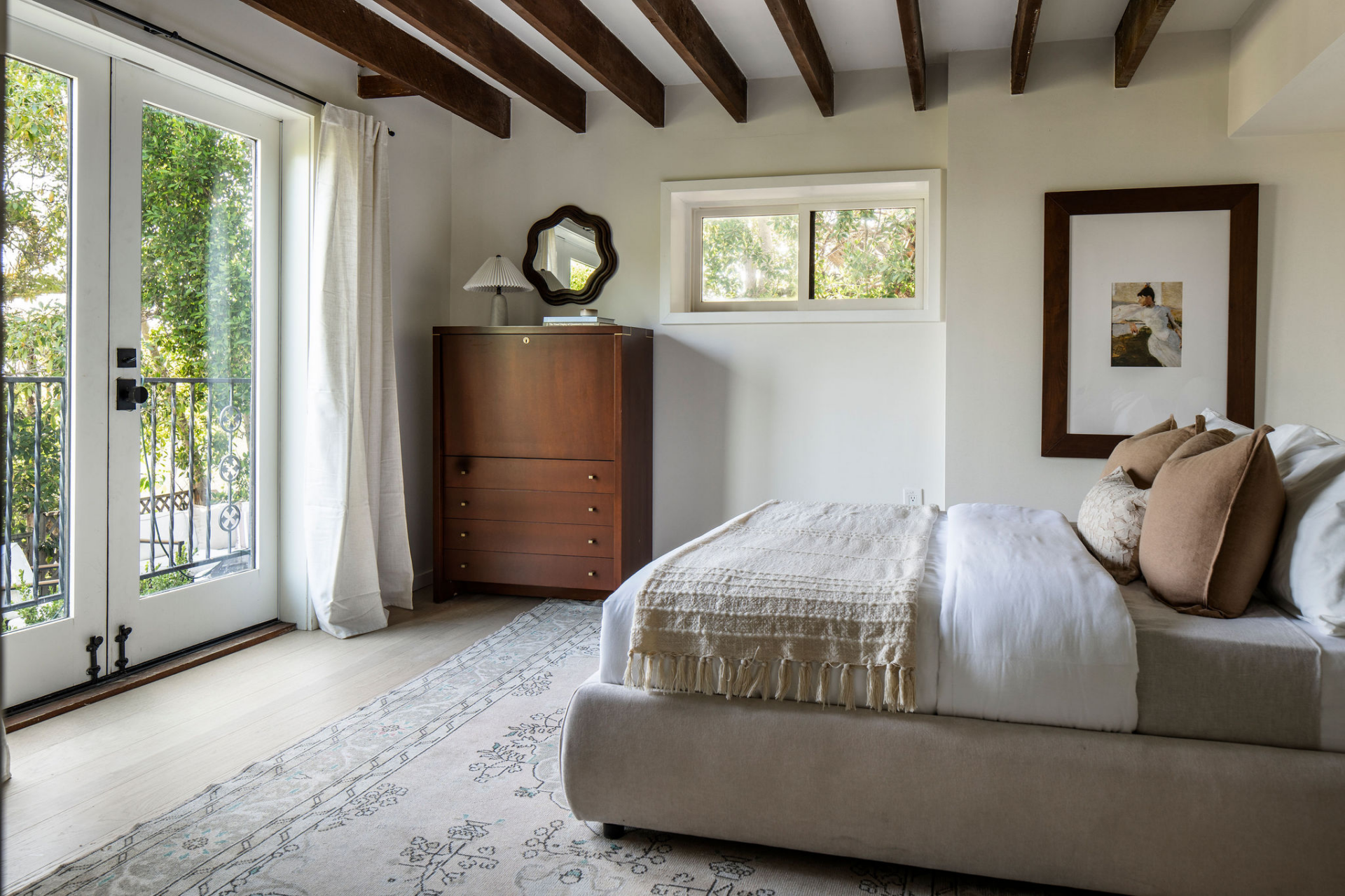 French doors open directly to a private outdoor balcony, while neutral furnishings, layered textiles, and classic wood furniture complete the space.