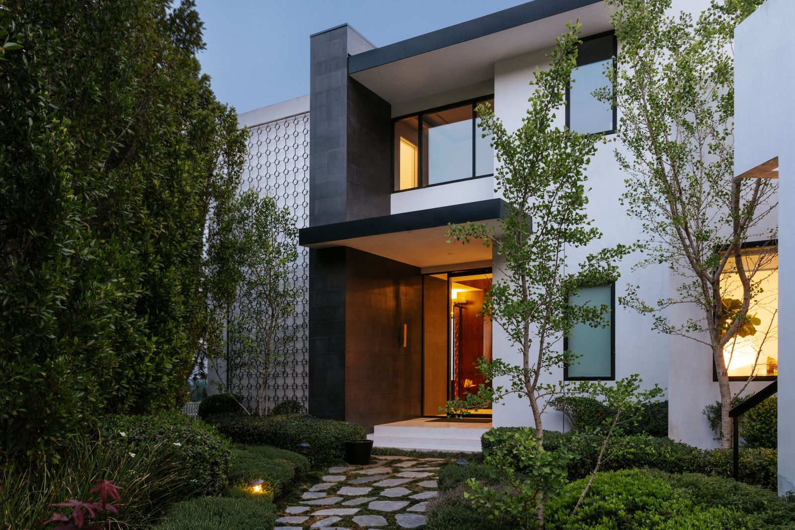 A winding stone pathway leads through manicured landscaping to the home's striking front entrance, where crisp white stucco is contrasted by charcoal stone and a decorative metal addition. 