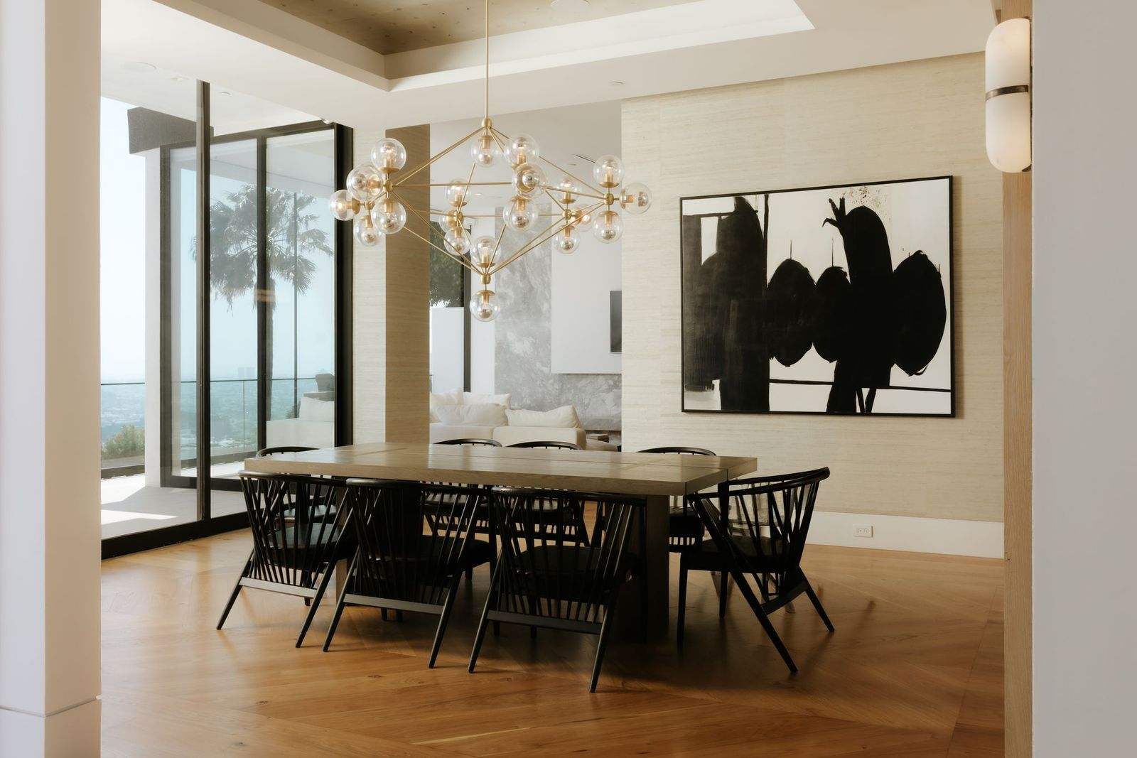 A long wood dining table is surrounded by black spindle-back chairs, while floor-to-ceiling glass draws in natural light.