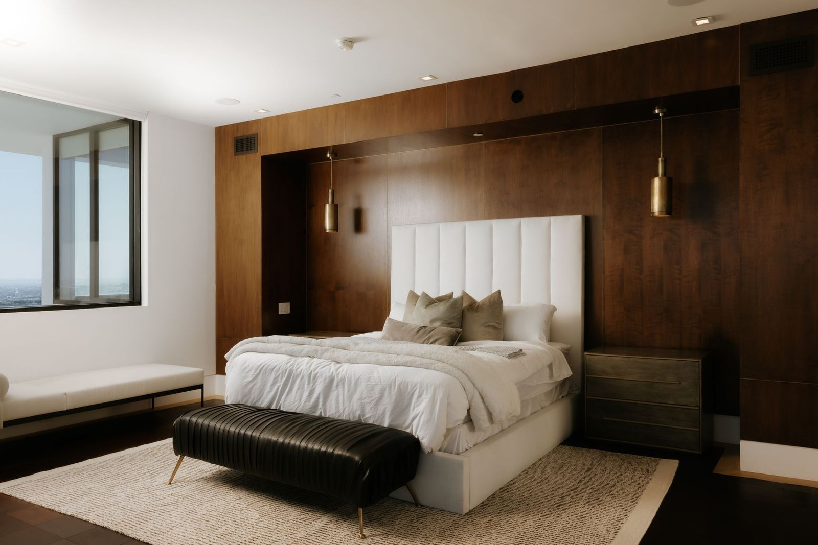 The bedroom is centered around a tall upholstered headboard set against a wall of rich walnut, complemented by minimalist furnishings and warm wood flooring. 