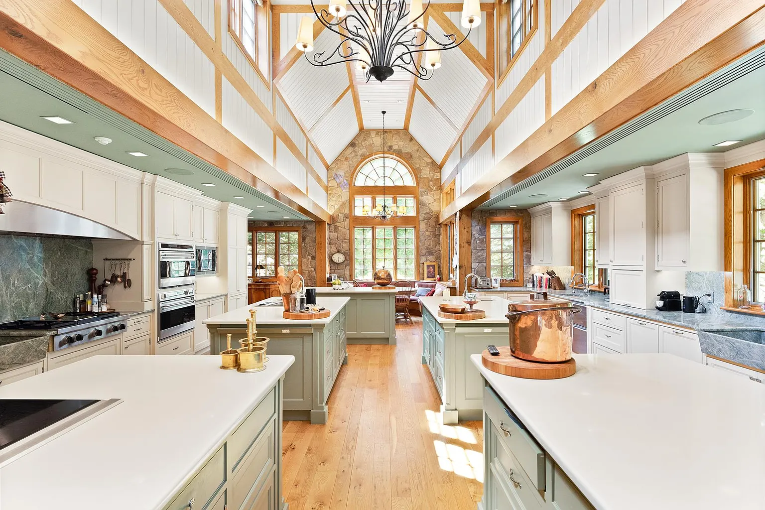 The commercial-grade kitchen is designed beneath a soaring vaulted ceiling in white tongue-and-groove paneling, exposed timber trusses, and oversized chandeliers. 