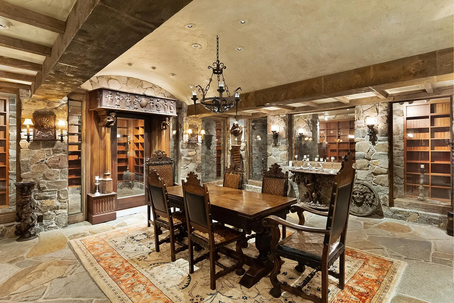 A long tasting table sits beneath wrought-iron chandeliers, while glass-lined storage rooms display extensive wine collections surrounding the space.