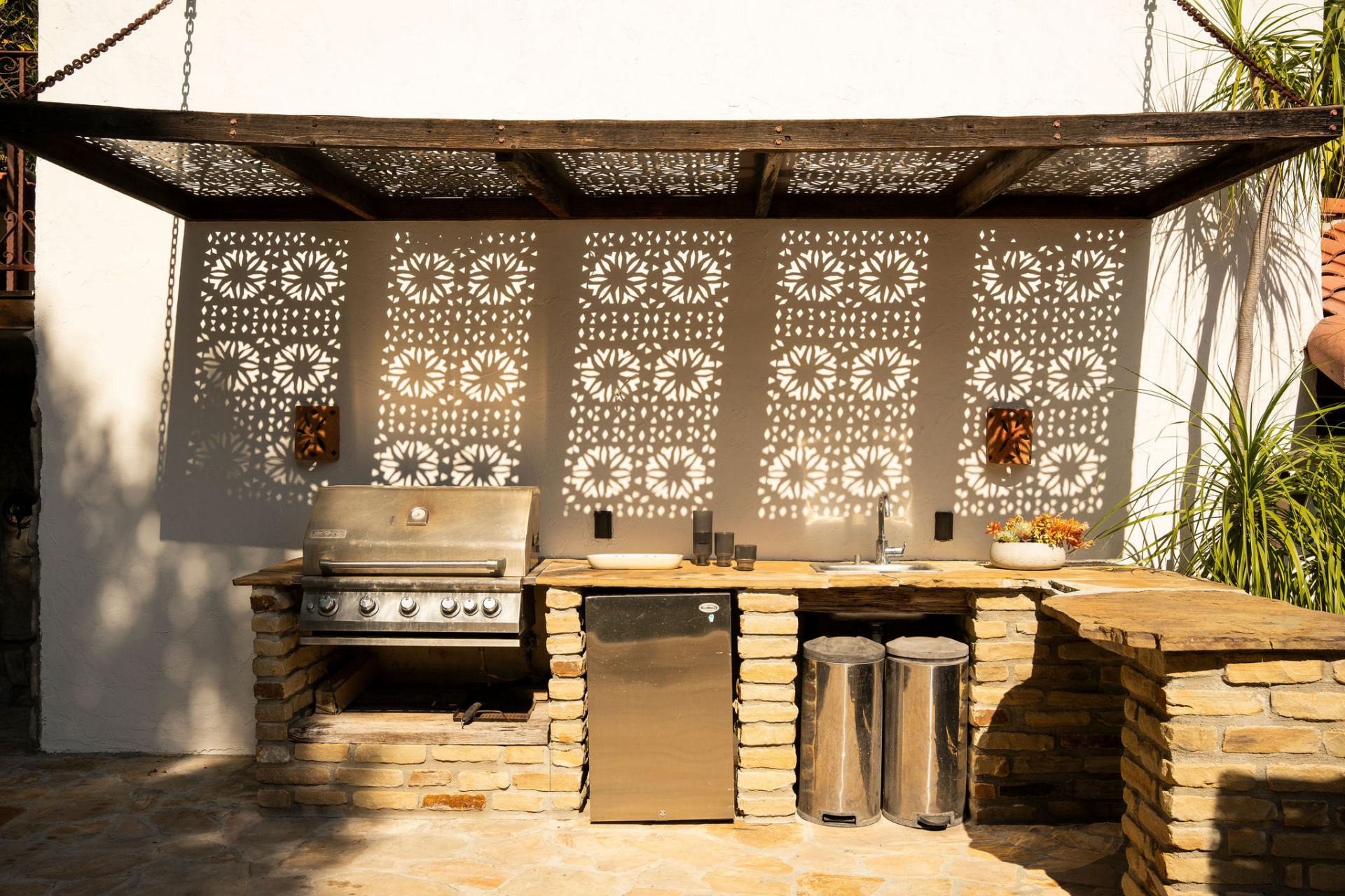 Stone counters, a built-in grill, under-counter refrigeration, and a sink provide everything needed for outdoor cooking, while decorative patterned screens cast shifting shadows across the space.