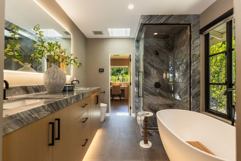A spacious walk-in shower is finished in dramatic gray stone, while a floating dual-sink vanity with stone countertops, backlit mirrors, and custom cabinetry sits across the room