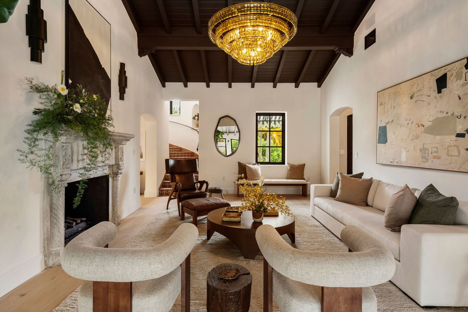 Wide-plank wood flooring runs throughout the space, beneath a seating arrangement of sofas, lounge chairs, and a round wood coffee table all positioned to face an original carved limestone fireplace.