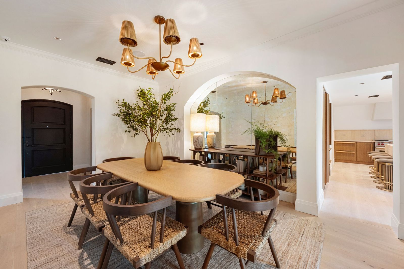 A large oval dining table sits at the center of the room with eight woven-seat chairs arranged around it, all positioned beneath a sculptural brass chandelier.