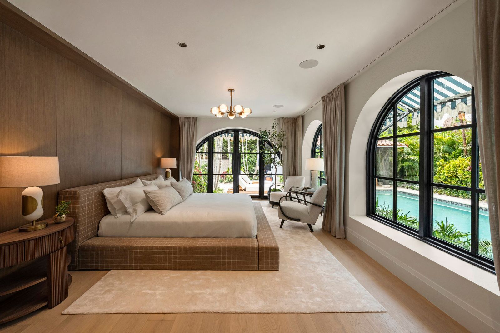 A full wall of warm wood paneling serves as the backdrop for the upholstered bed, while matching arched windows and doors frame views of the landscaped backyard.