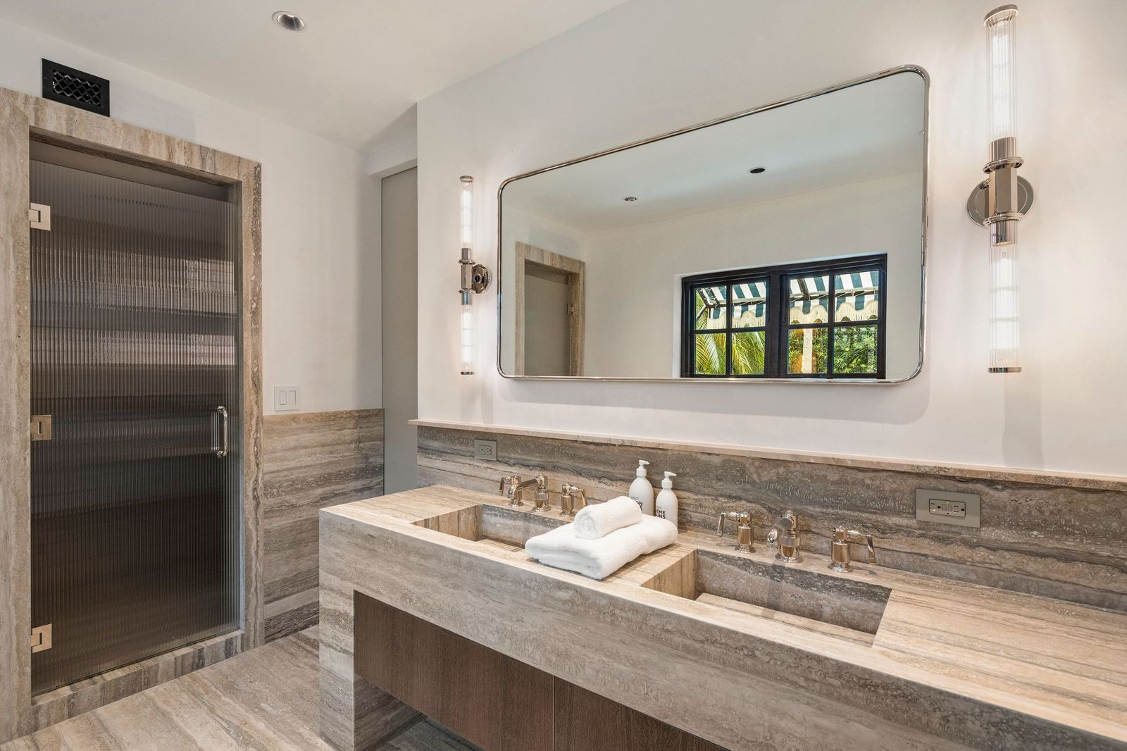A long floating dual-sink vanity sits beneath a wide framed mirror framed by vertical sconces, while a glass-enclosed walk-in shower finished in matching stone.