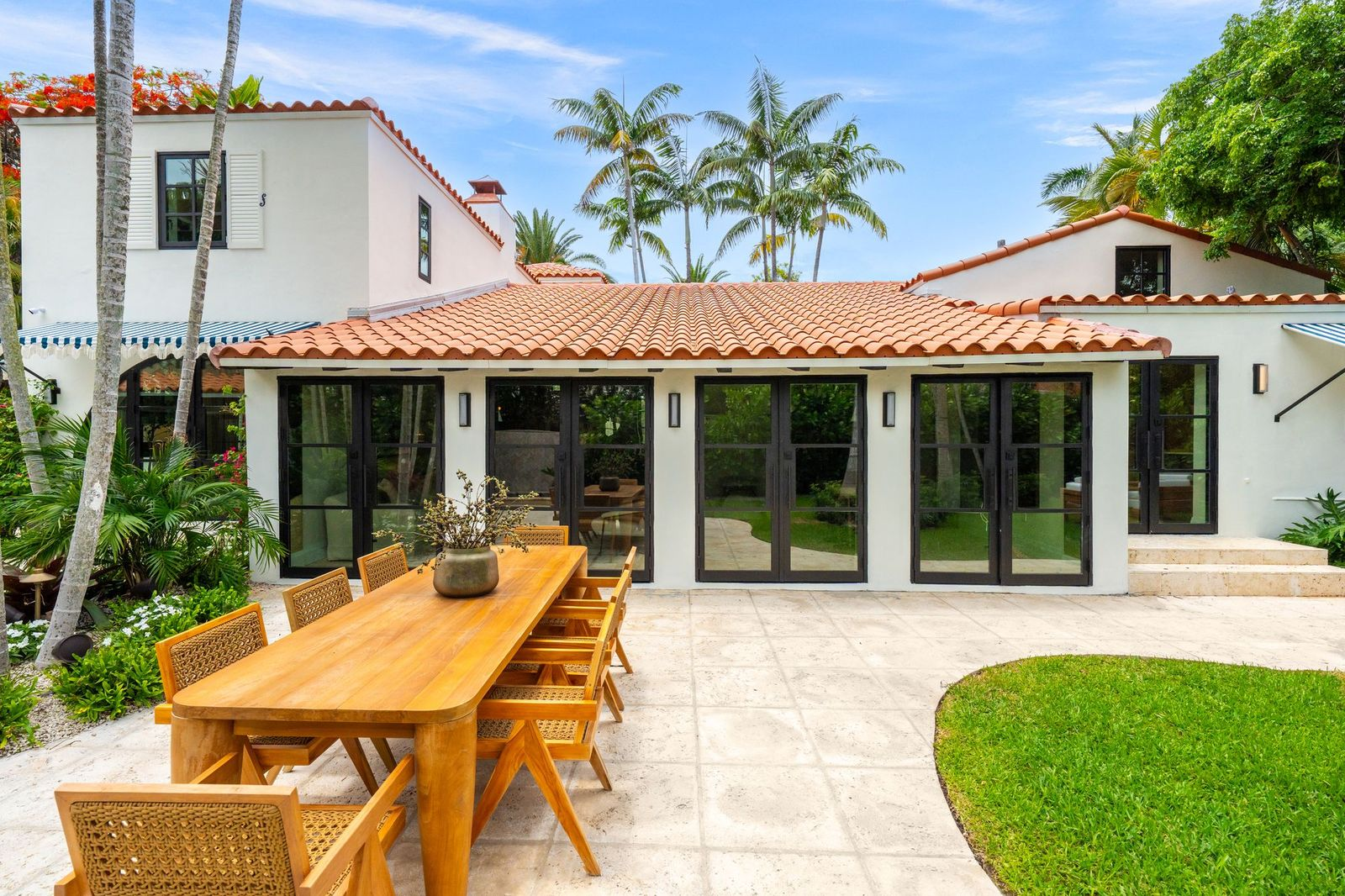 A large stone patio has a long teak dining table with seating for eight placed atop it.