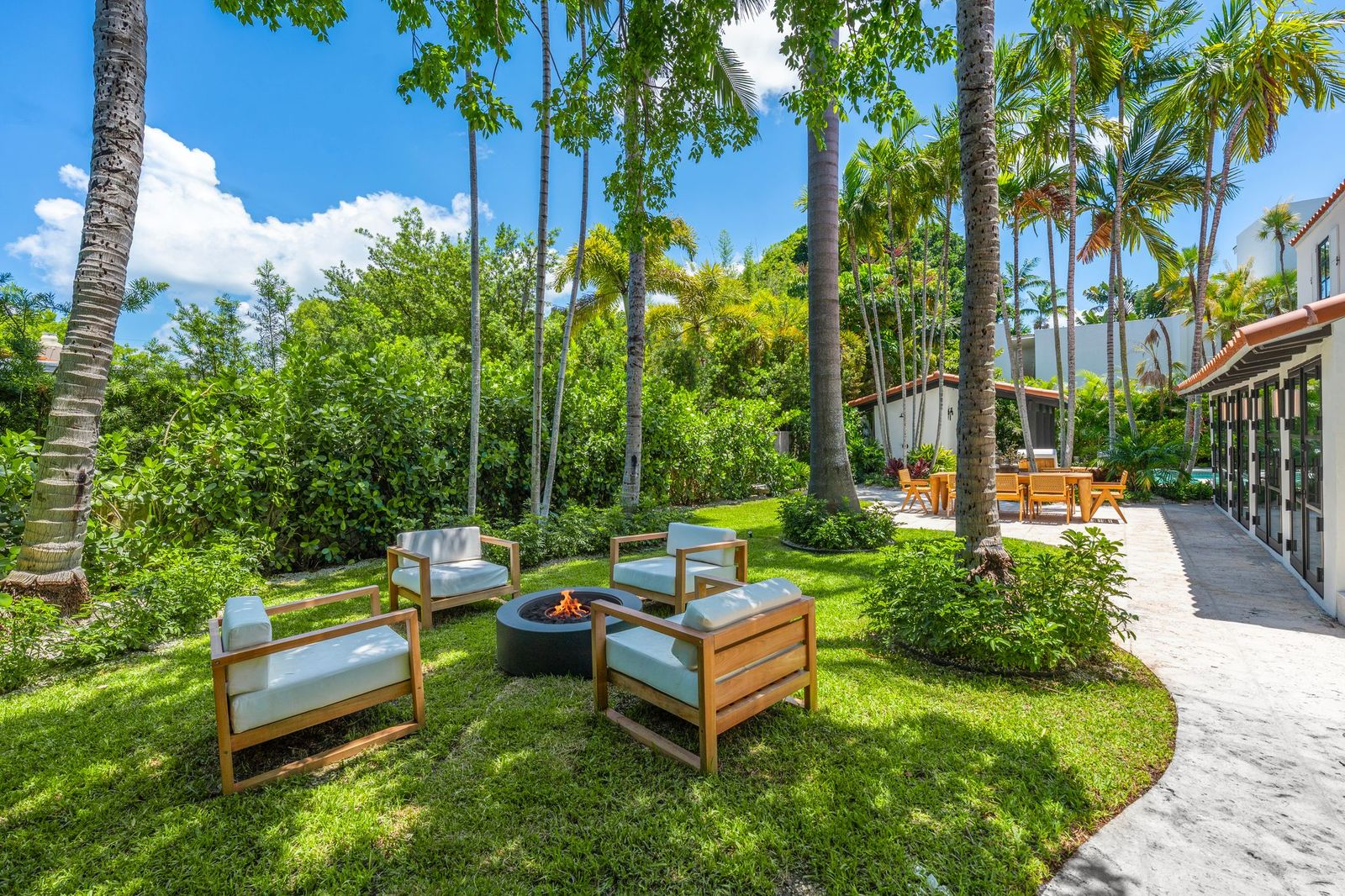A  curved stone path winds through the lawn to a circular fire pit lounge tucked beneath a canopy of palm trees. 