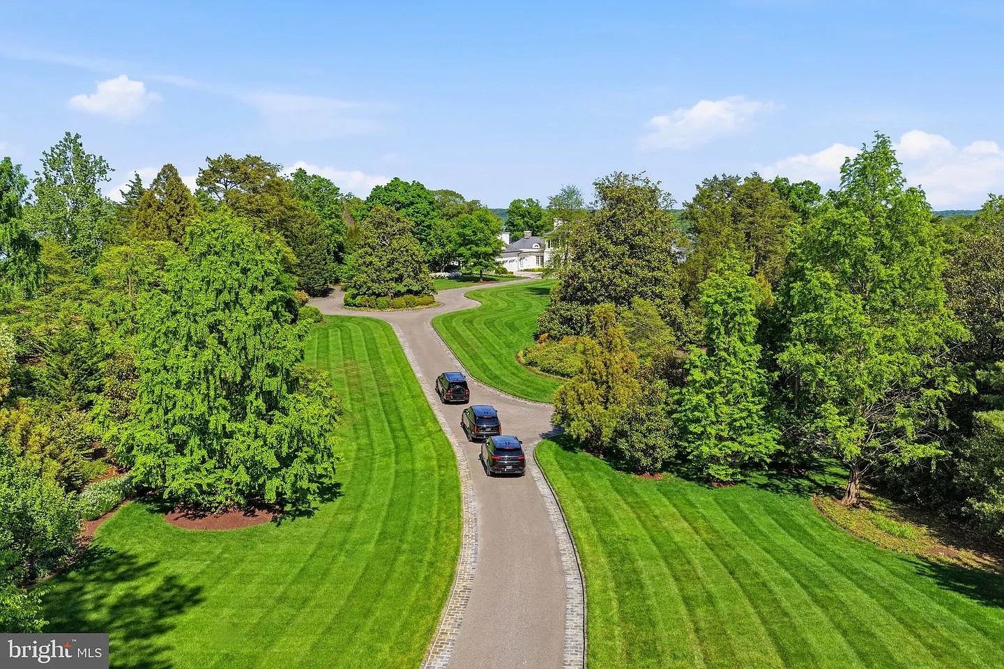 A winding stone-lined driveway cuts through more than 16 acres of grounds, bordered by trees, sculpted hedges, and expansive lawns striped with precise mowing patterns. 