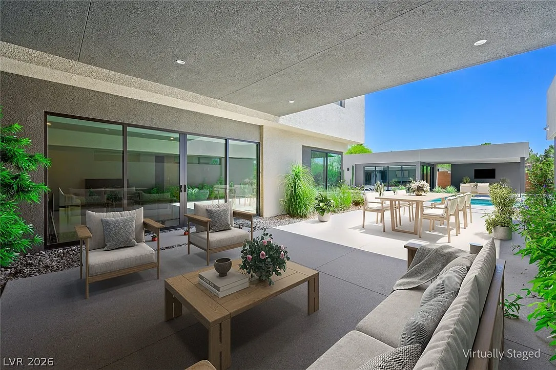 One end is arranged as an outdoor living room beneath the shade of the overhang, while farther along, the patio opens to a large dining table positioned for outdoor meals.