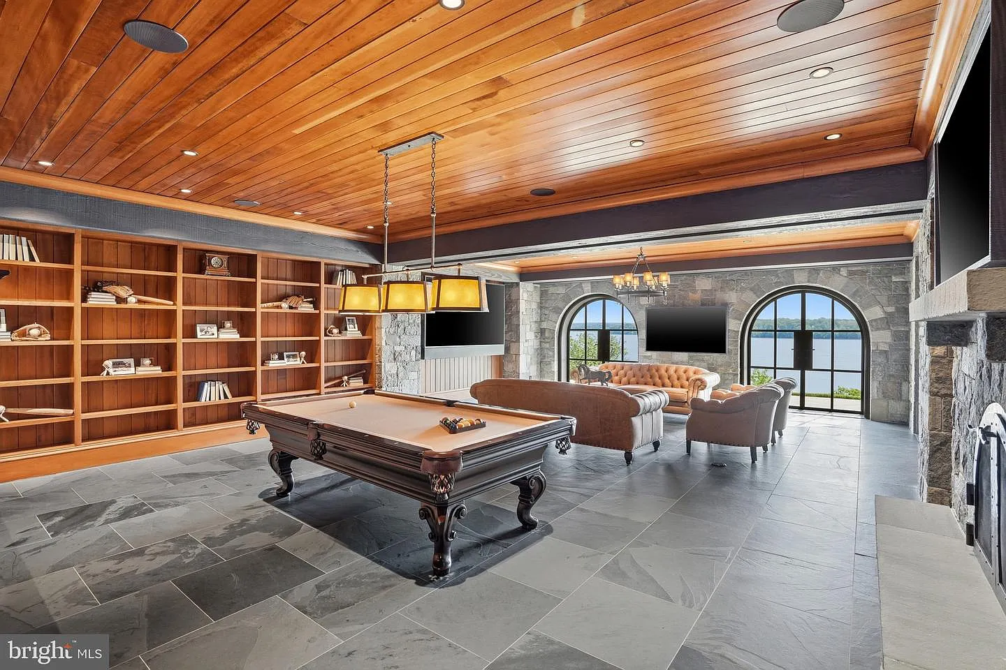 A billiards lounge with a full-size pool table, floor-to-ceiling shelving, and a seating area positioned near the windows.