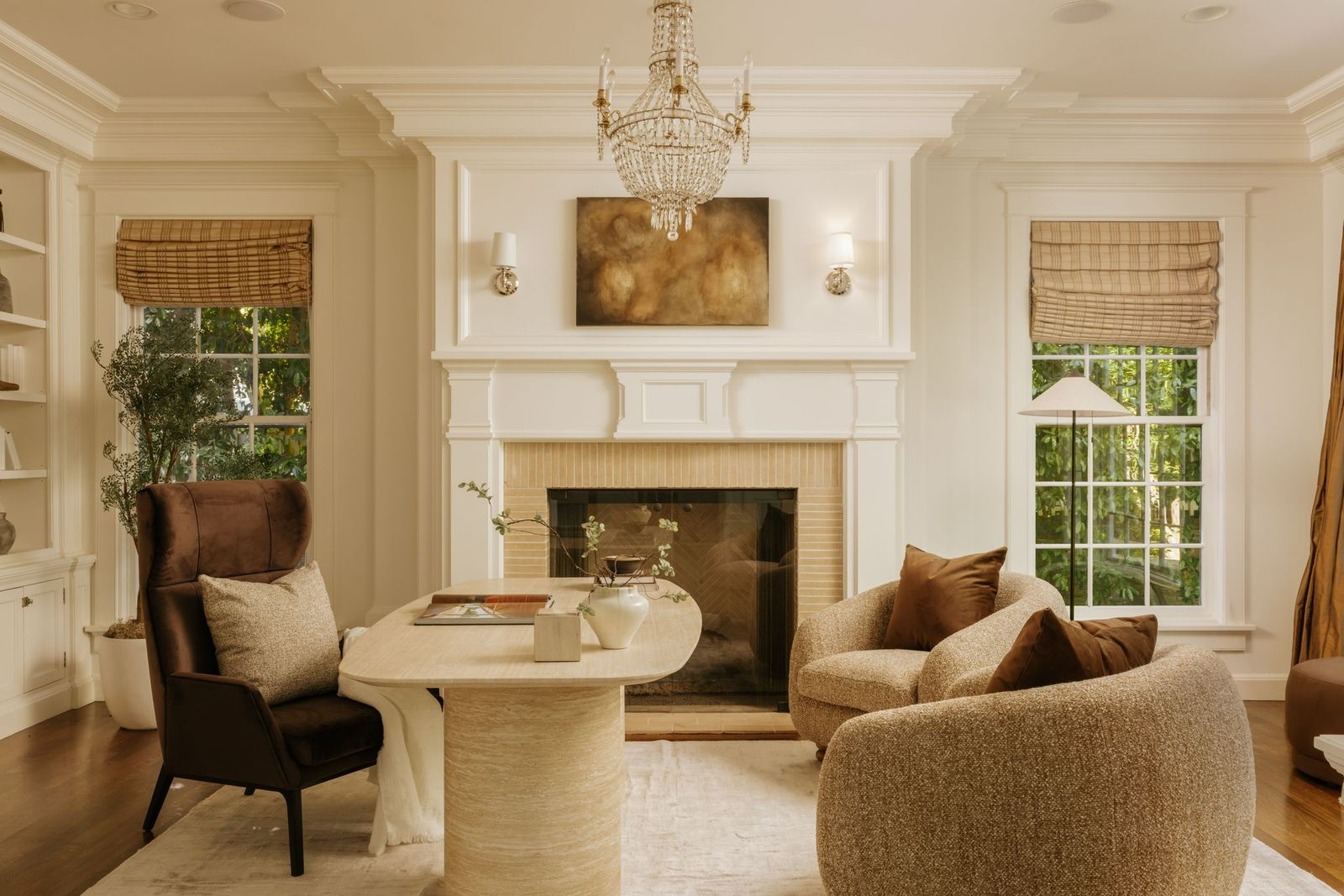 A fireplace serves as the focal point behind the sculptural desk, surrounded by paneled walls and symmetrical windows dressed in woven Roman shades.