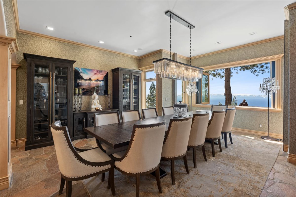 A long dining table that sits beneath a long crystal chandelier, with large picture windows framing Lake Tahoe as the backdrop. 