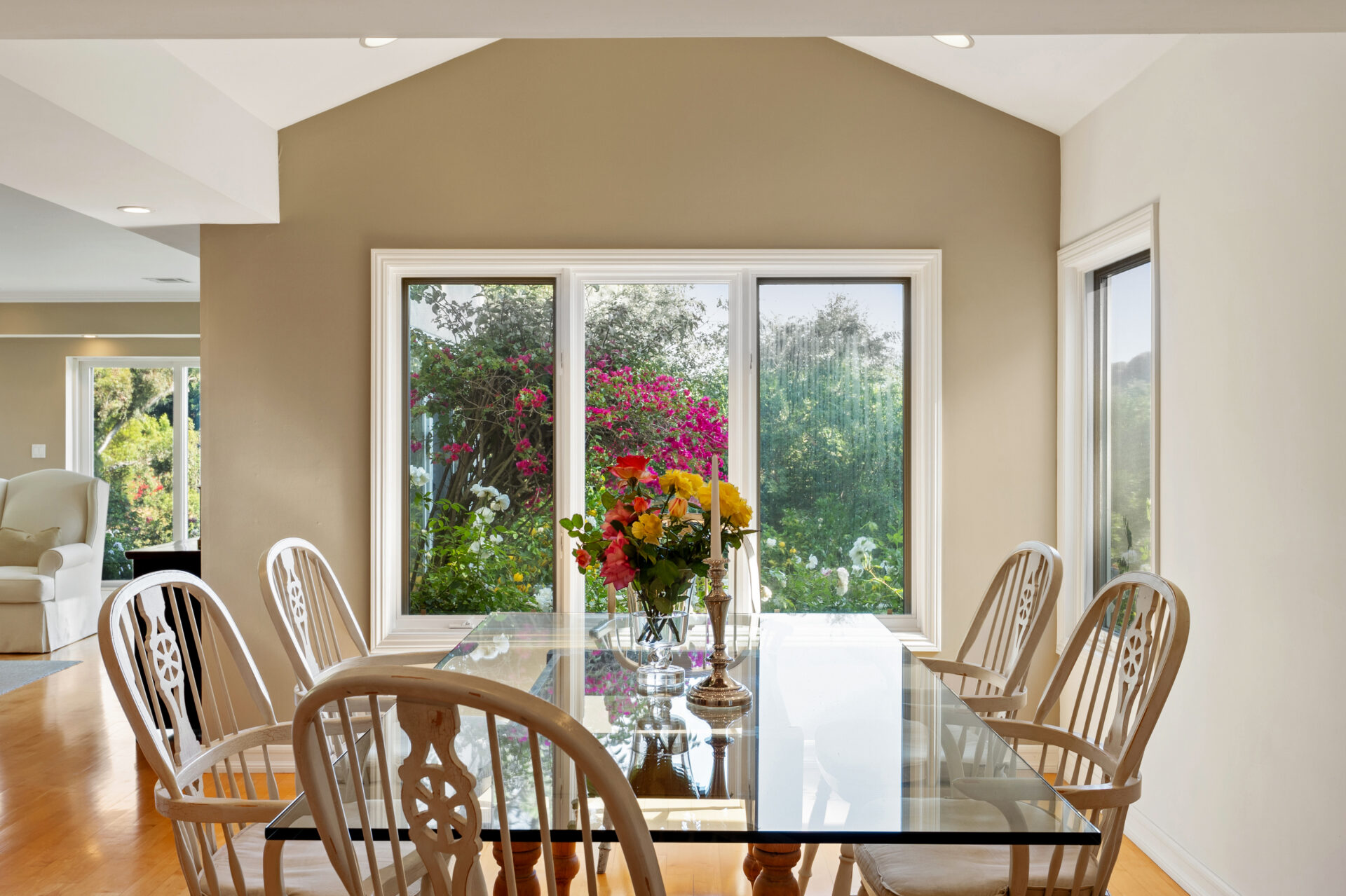 A glass-top dining table surrounded by white spindle-back chairs looking out a large window.