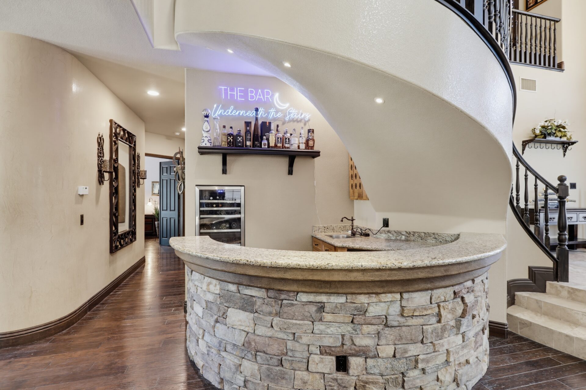 Tucked beneath the staircase, a curved stone bar with granite countertops, a built-in wine refrigerator, and a sink.