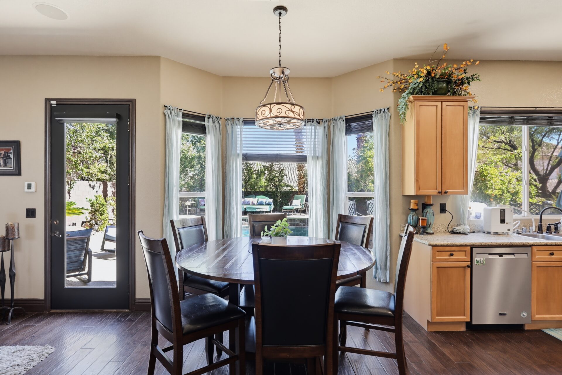 A round wood table sits beneath an iron light fixture, while a bay of windows fills the room with natural light and frames views of the backyard pool and landscaping.