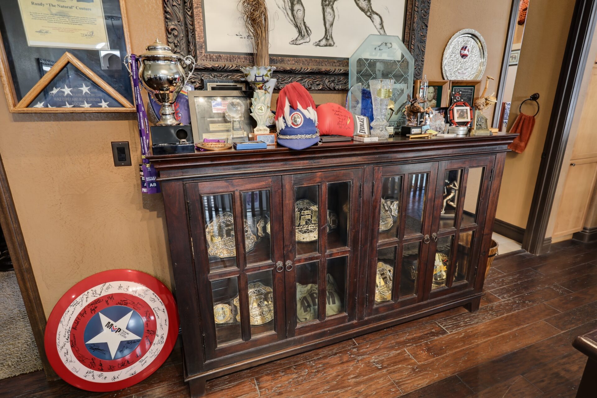 Couture's career memorabilia in a glass front cabinet, including championship belts, trophies, awards, and signed gloves.