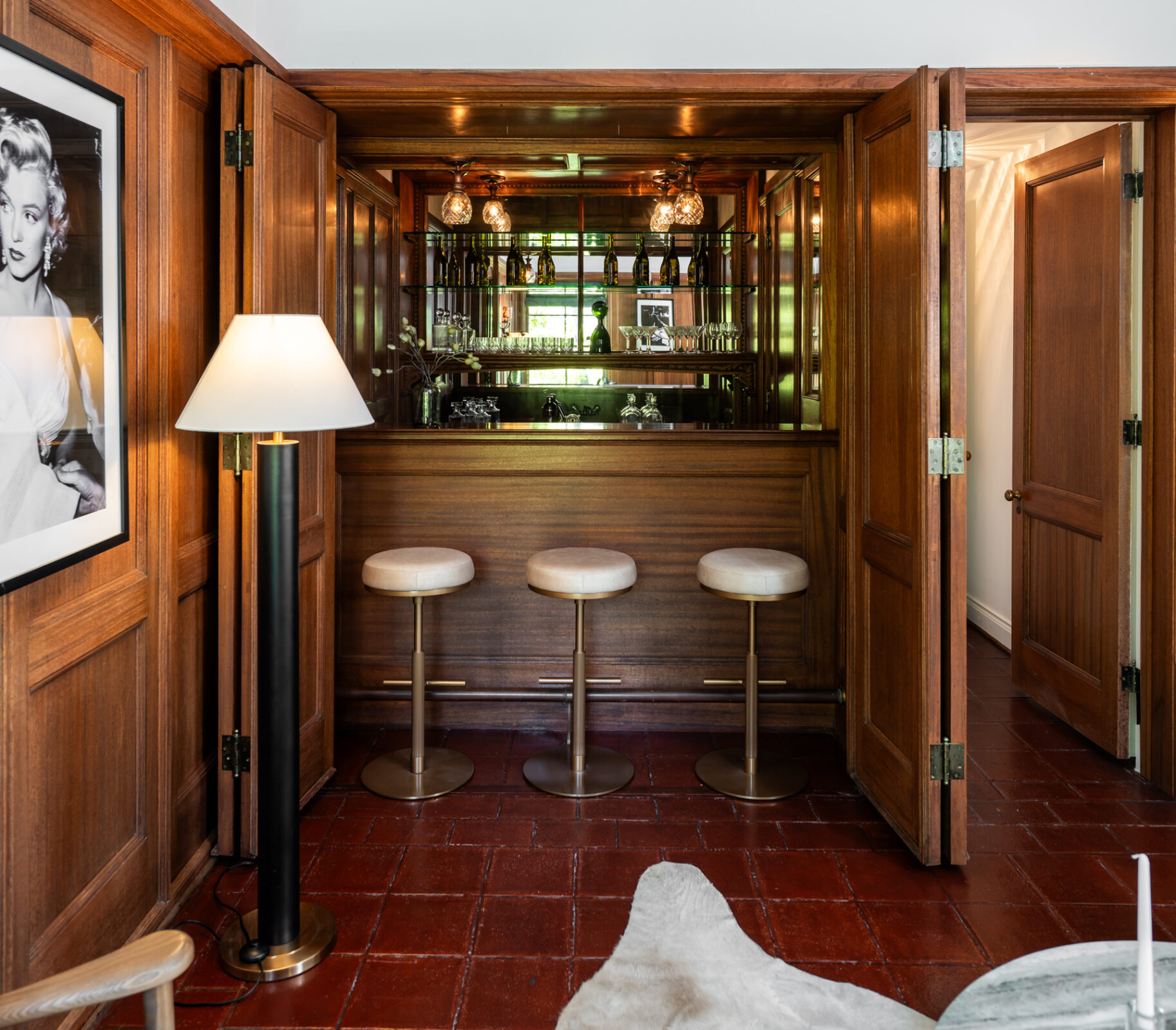 Foldable doors conceal a fully equipped wet bar, which opens to reveal mirrored shelving, glass display racks, and bartop seating for three.