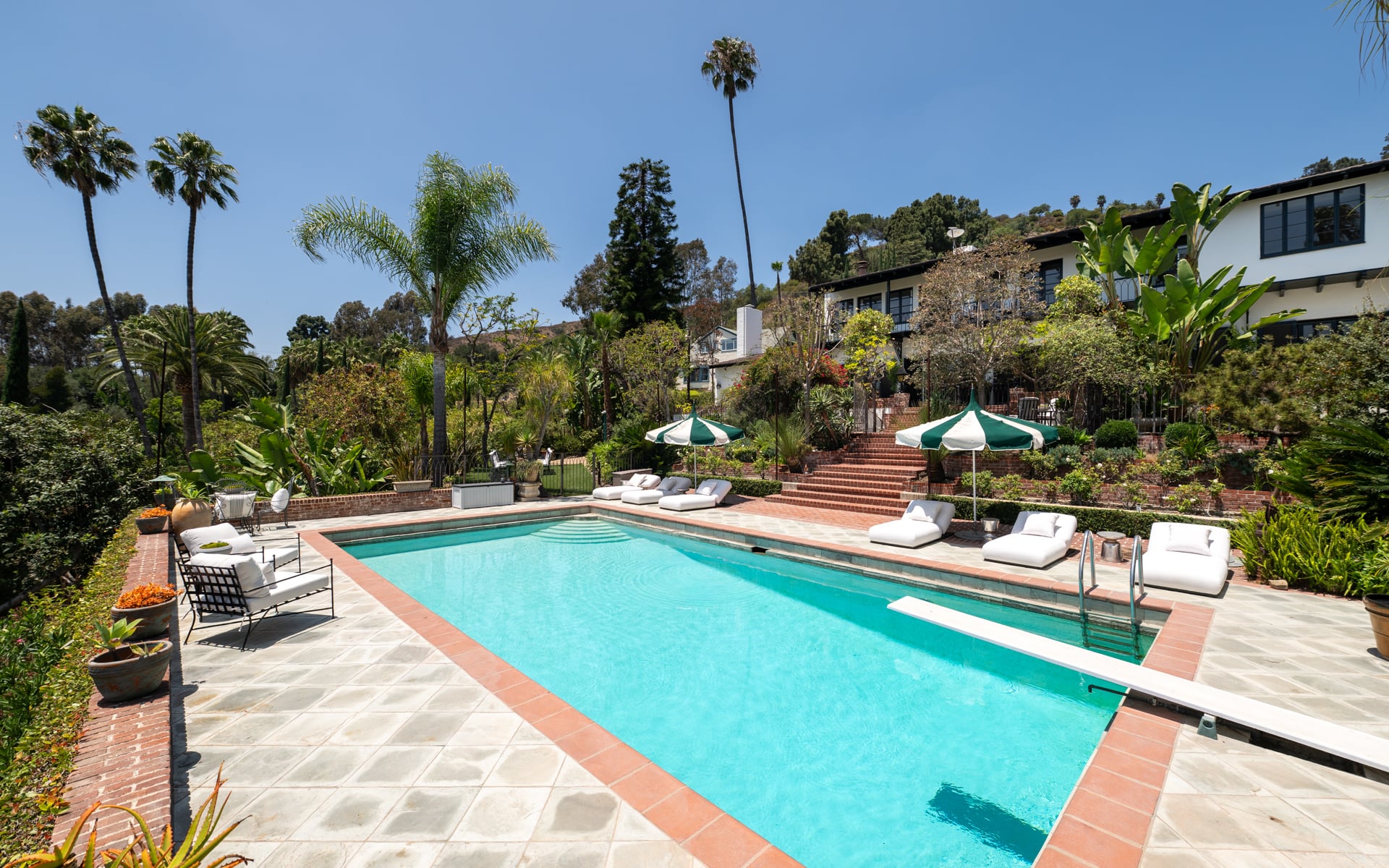 At the center of the backyard, the rectangular swimming pool is framed by brick terraces, hedges, and lounge chairs that line the pool deck.