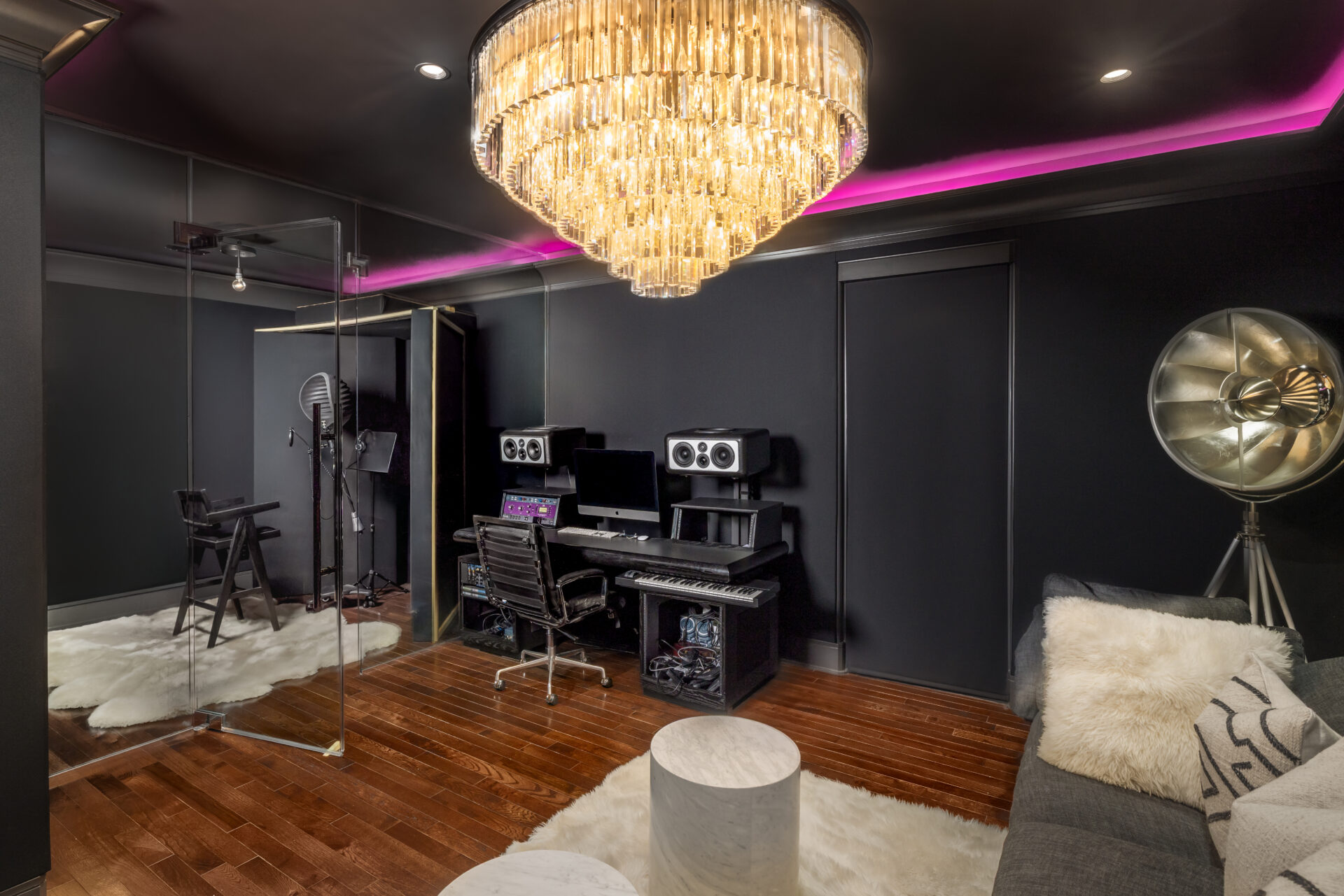 A mixing station with studio monitors and keyboard sits beside a glass-enclosed vocal booth, while a sofa and small marble tables create a separate listening area along one side of the room.