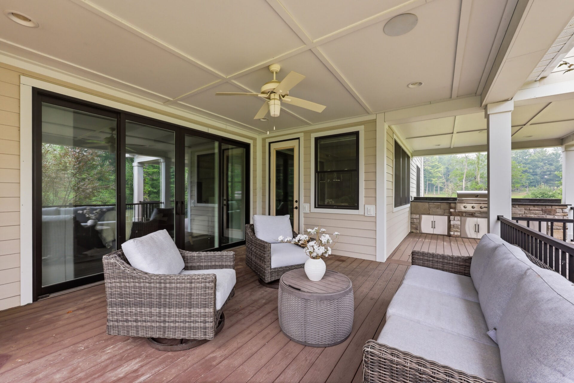 An outdoor living room is arranged beneath a finished ceiling with recessed lighting and a ceiling fan, while a built-in grilling station sits at one end overlooking the private, tree-lined backyard.