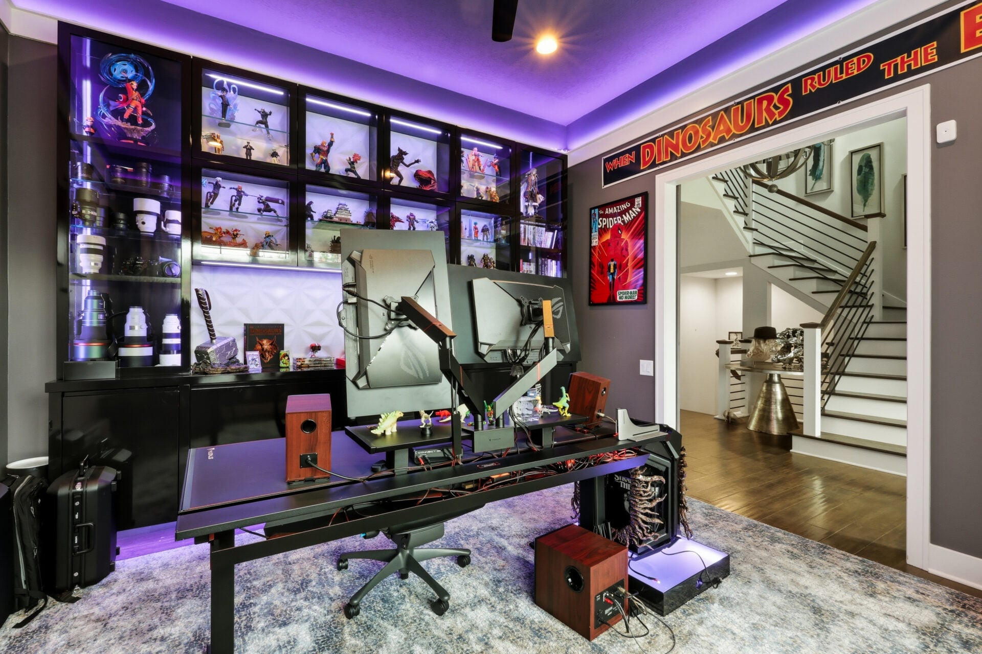 Across from the staircase, a pair of sliding barn doors opens into Garrett's dedicated gaming room, a space that reflects one of his biggest interests away from the football field. A custom workstation sits at the center with multiple monitors, while floor-to-ceiling display cabinets line one wall, showcasing an extensive collection of superhero figures, collectibles, and camera gear illuminated by integrated lighting. Purple LED accent lighting wraps the ceiling, comic book artwork decorates the walls, and themed memorabilia throughout the room gives the space a personalized, immersive atmosphere designed for gaming and streaming.